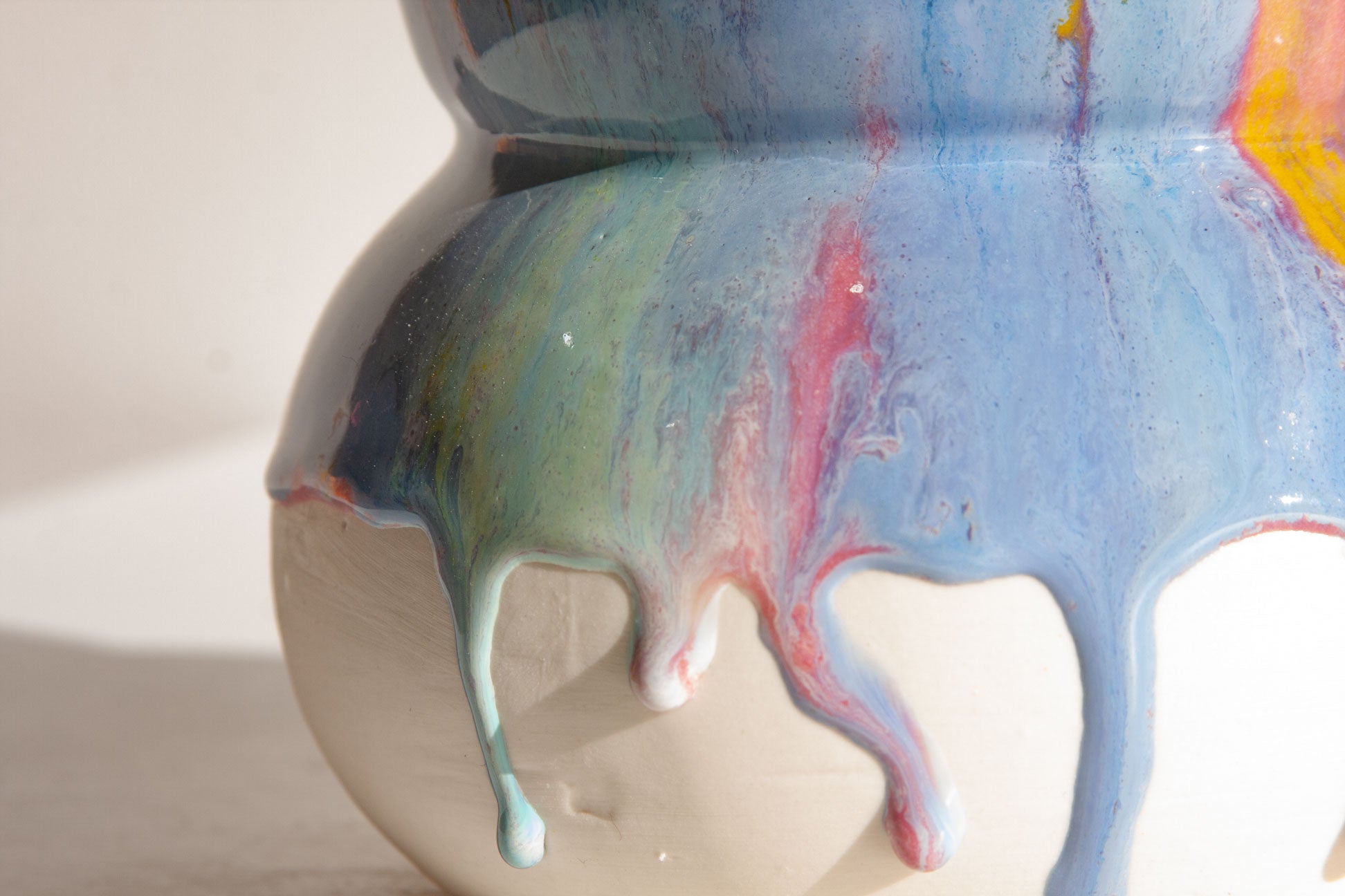 Close-up of a ceramic pot or vase with a cream-colored base and dripping multicolor glaze featuring blue, pink, green, and yellow swirls.