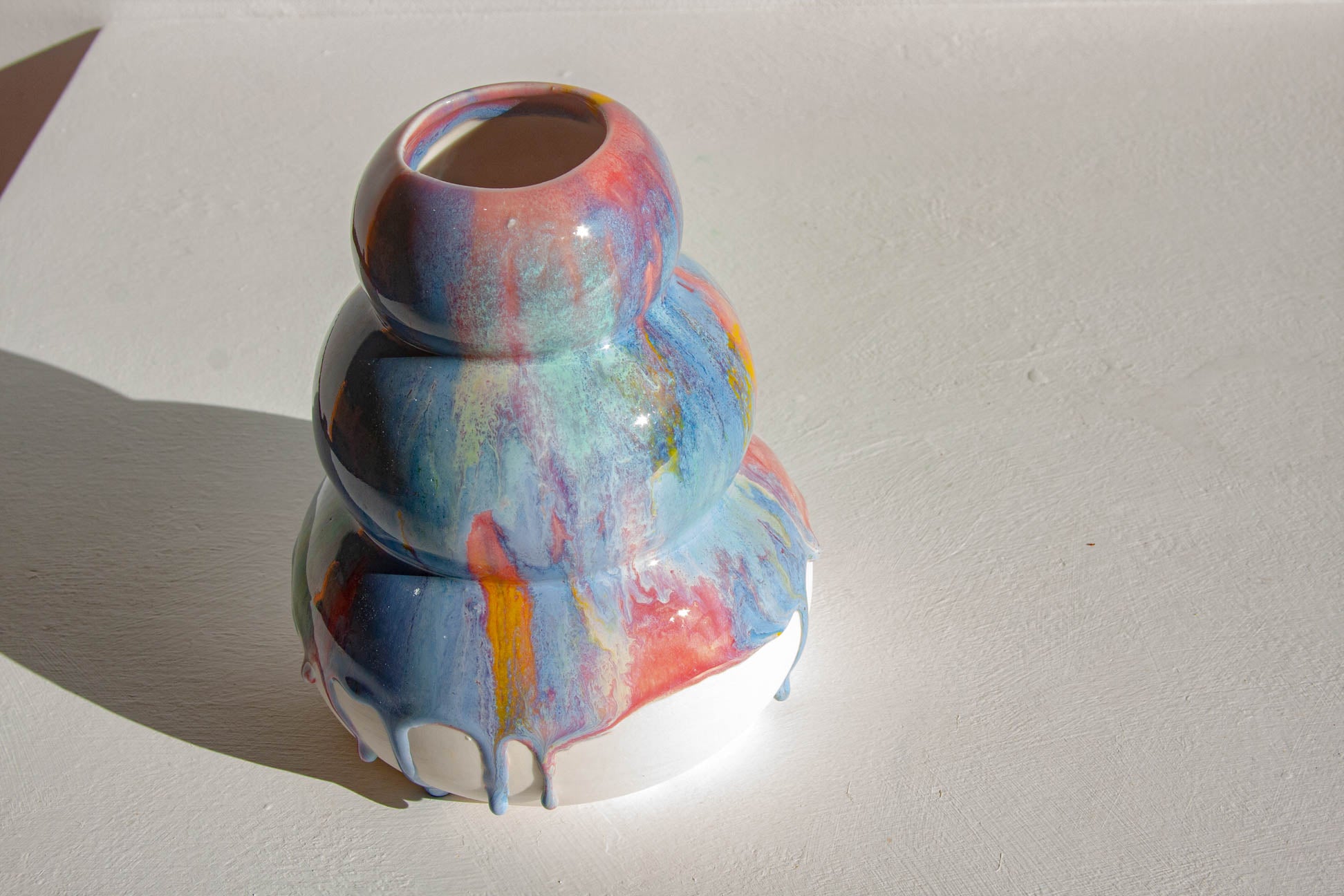 A sculptural ceramic vase with a dripping paint effect, featuring a marbled pattern of blue, pink, orange, and white on a white base, photographed on a white surface with a strong shadow.