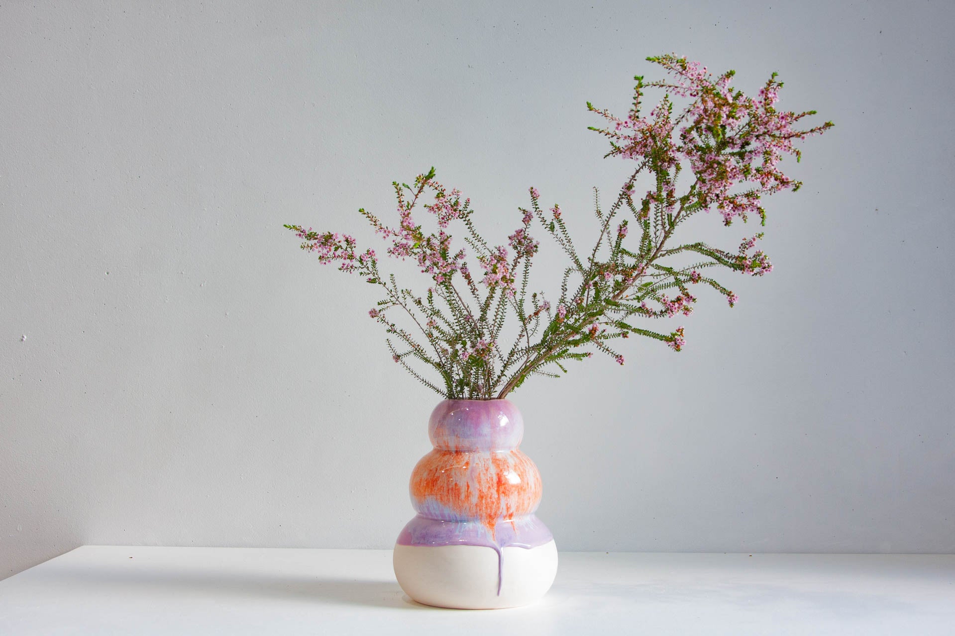 A small ceramic vase with a stacked, rounded design in white, orange, and lavender, holding delicate pink heather stems against a plain light grey wall and white surface.