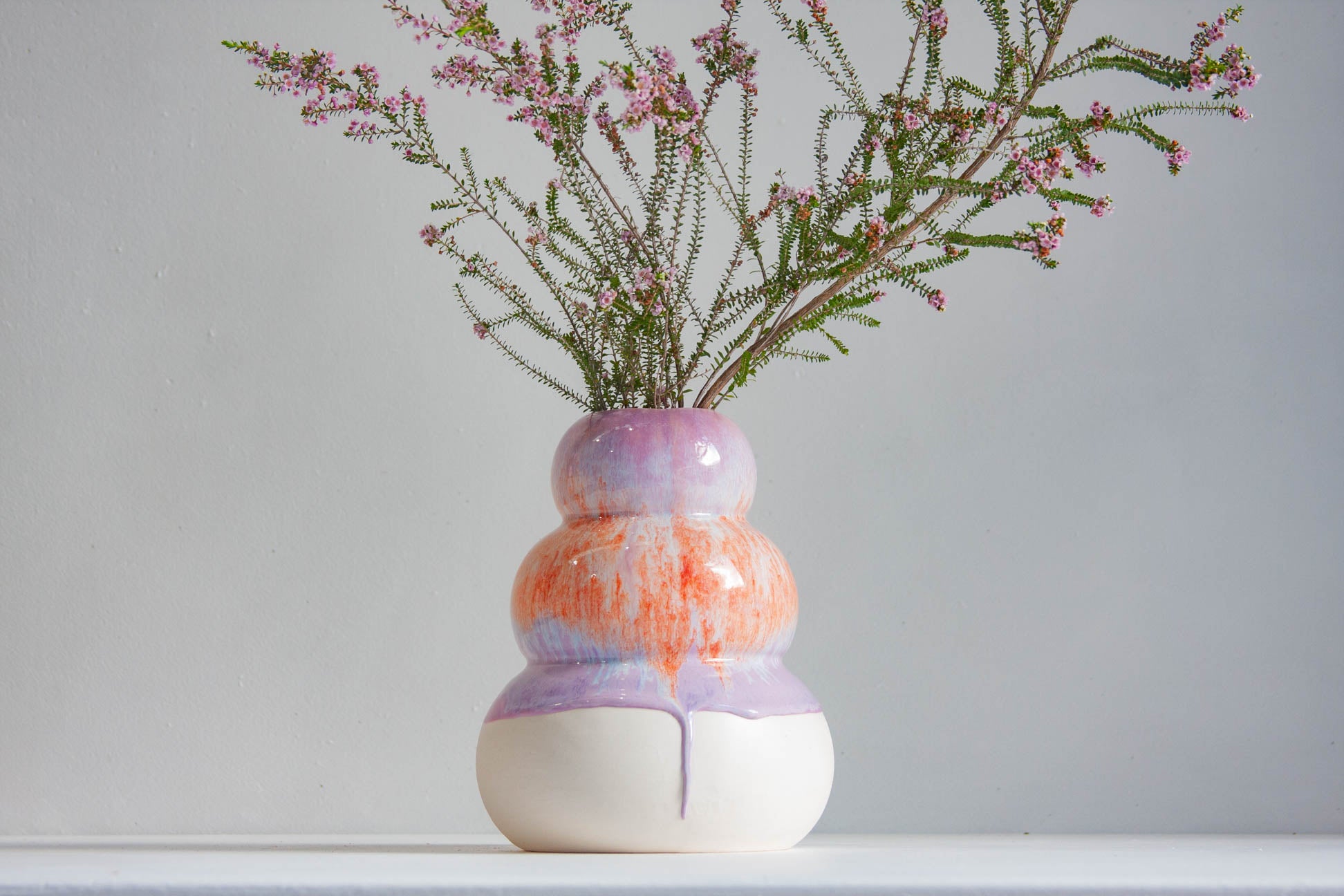 A ceramic vase with a stacked, rounded design in white, orange, and lavender purple, holding delicate pink heather stems against a neutral light grey background.