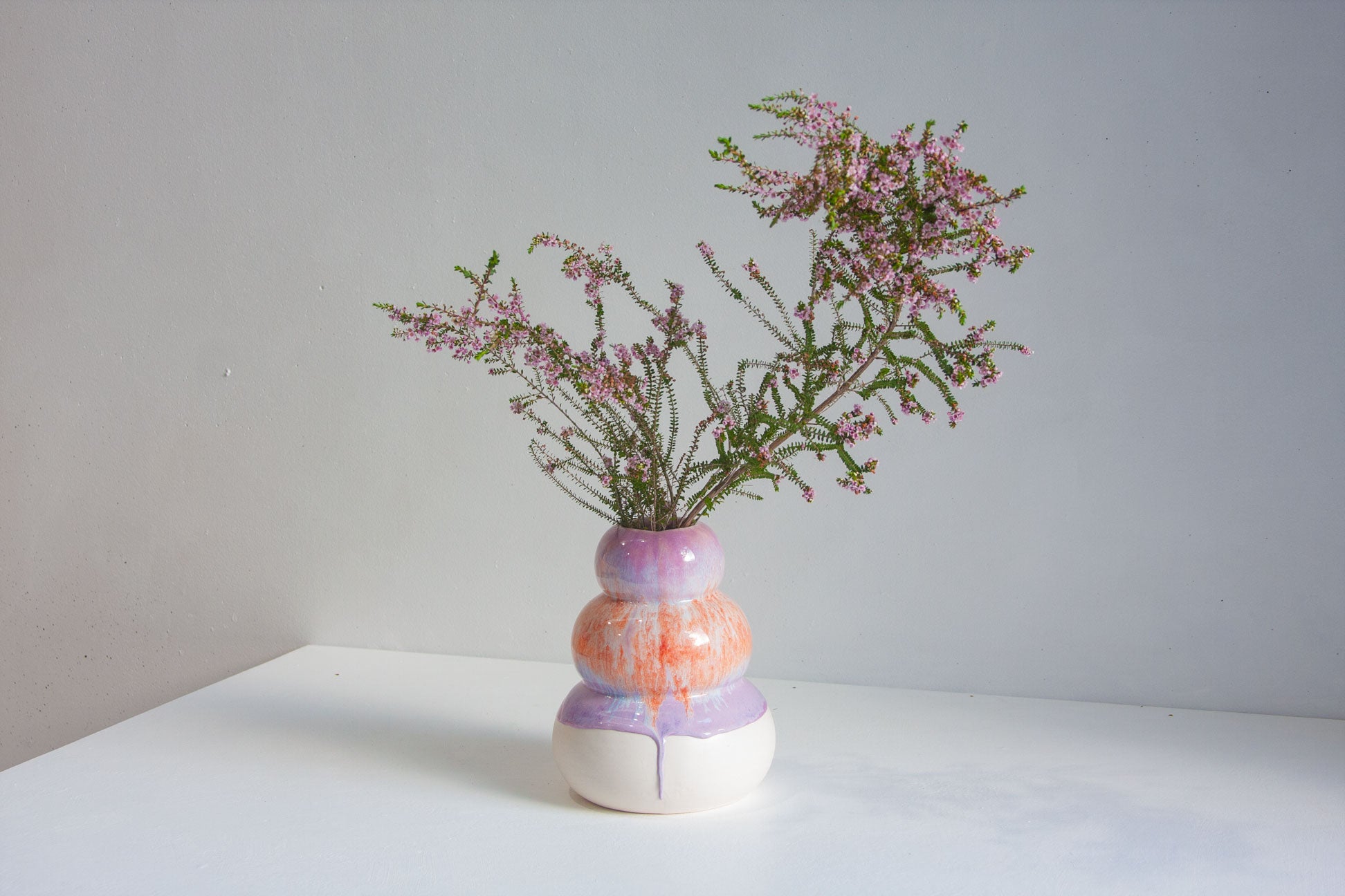 A small stacked ceramic vase with a glossy marbled finish in purple, orange, and white, holding dried pink heather stems, set on a white surface against a light grey wall.