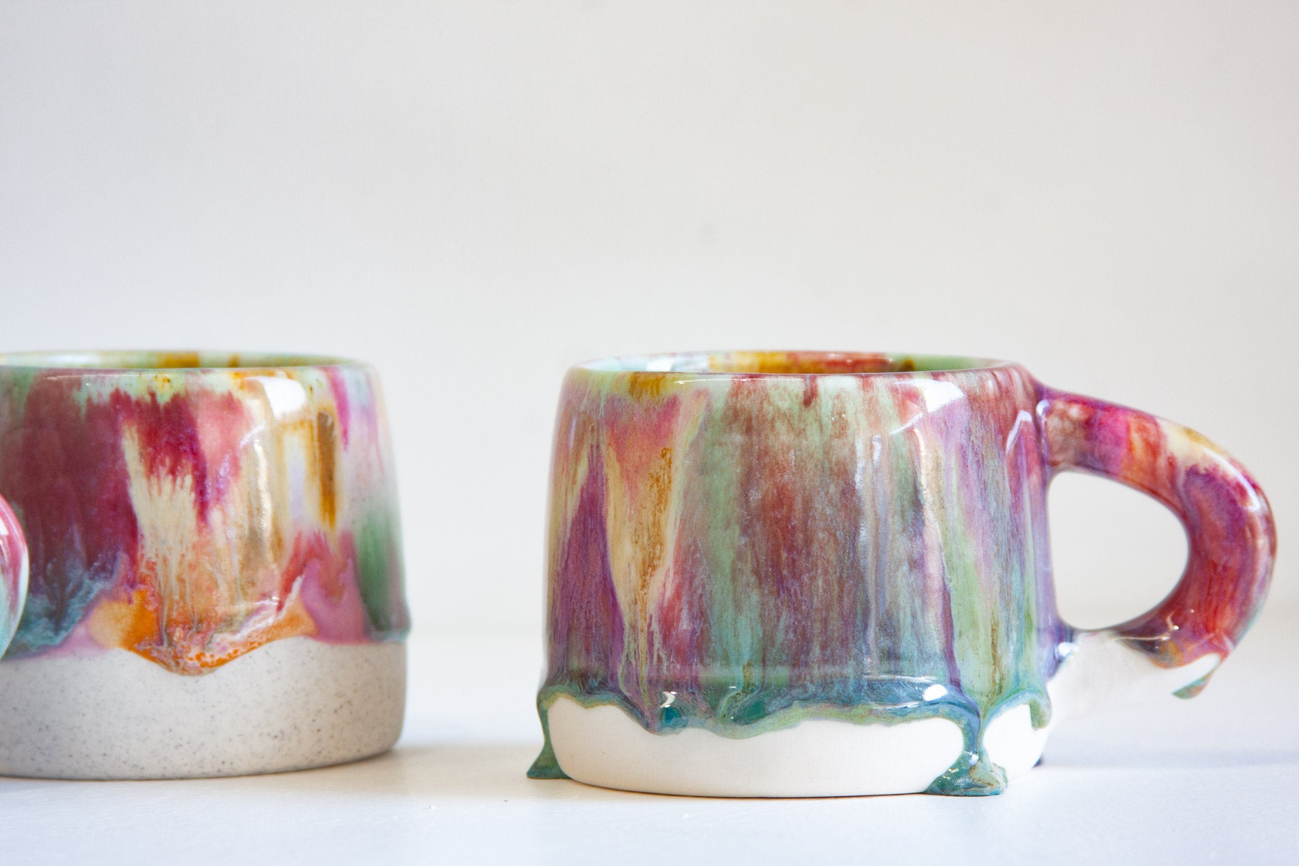 Two handmade ceramic mugs with a colorful drip glaze pattern, featuring pink, purple, green, and orange swirls over a white base, set against a plain white background.