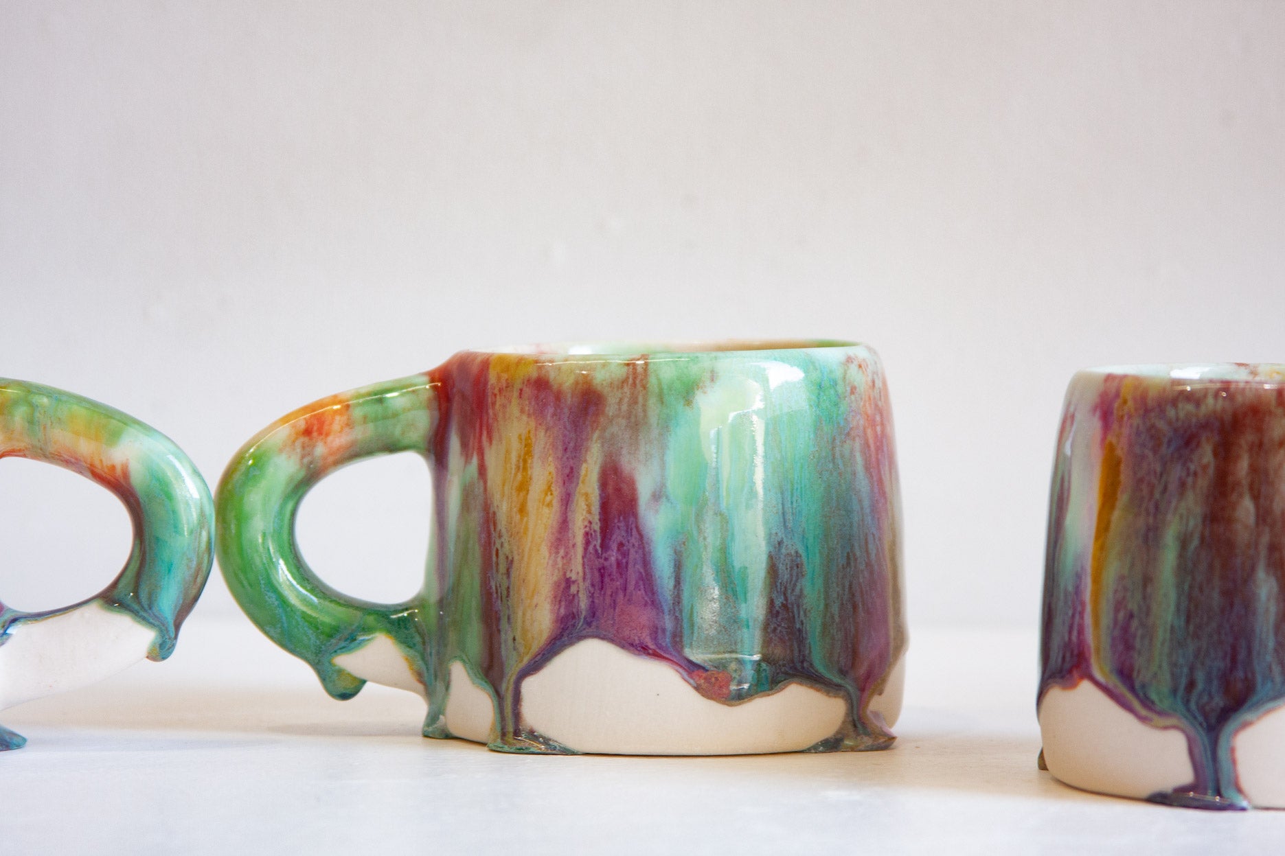 Three ceramic mugs with a glossy drip glaze finish, featuring a multicolor blend of green, purple, orange, and brown on a white base, set against a plain light background.