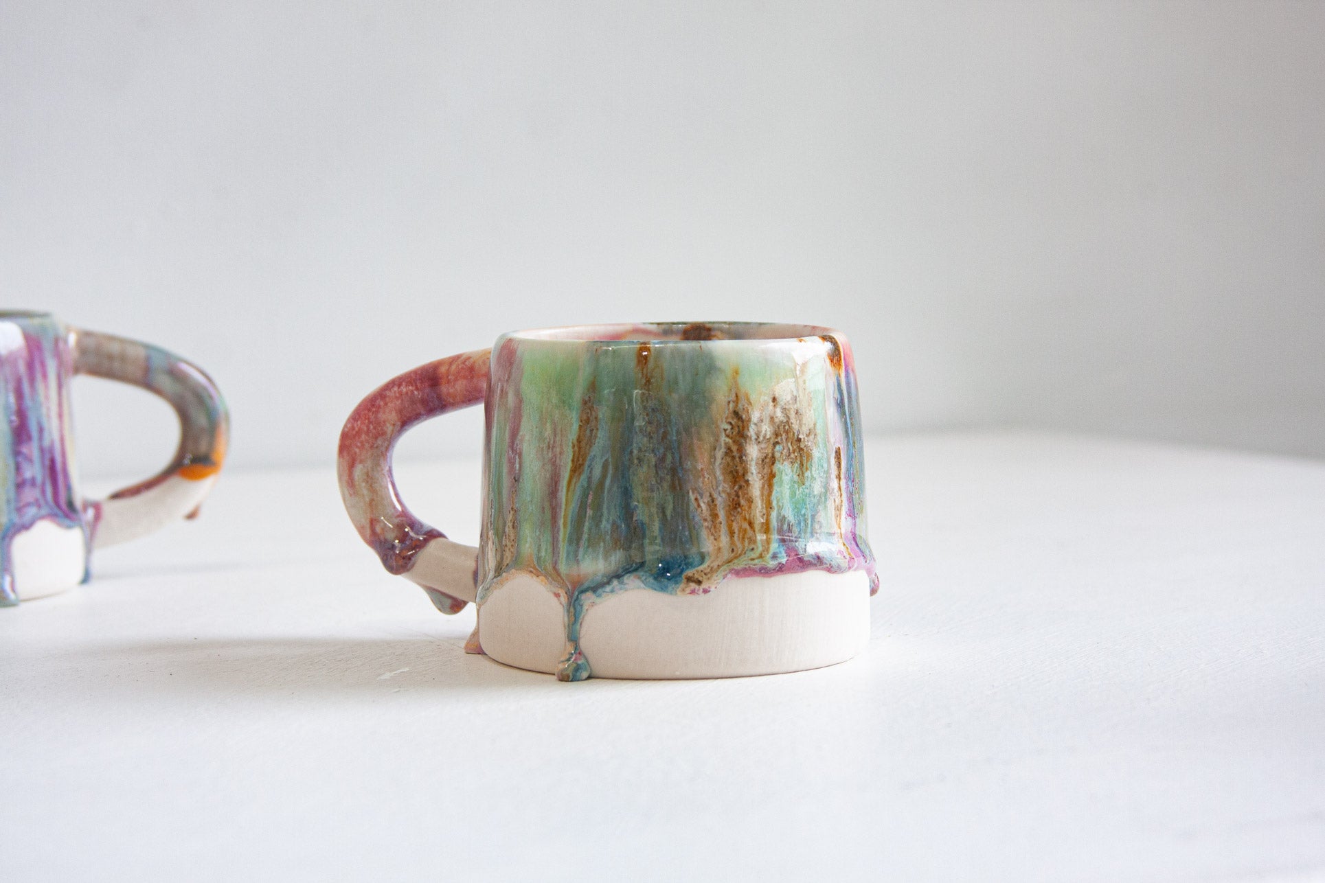 A handmade ceramic mug with its handle to the left, the glaze running in green, brown and pink streaks down to the bare white clay base.