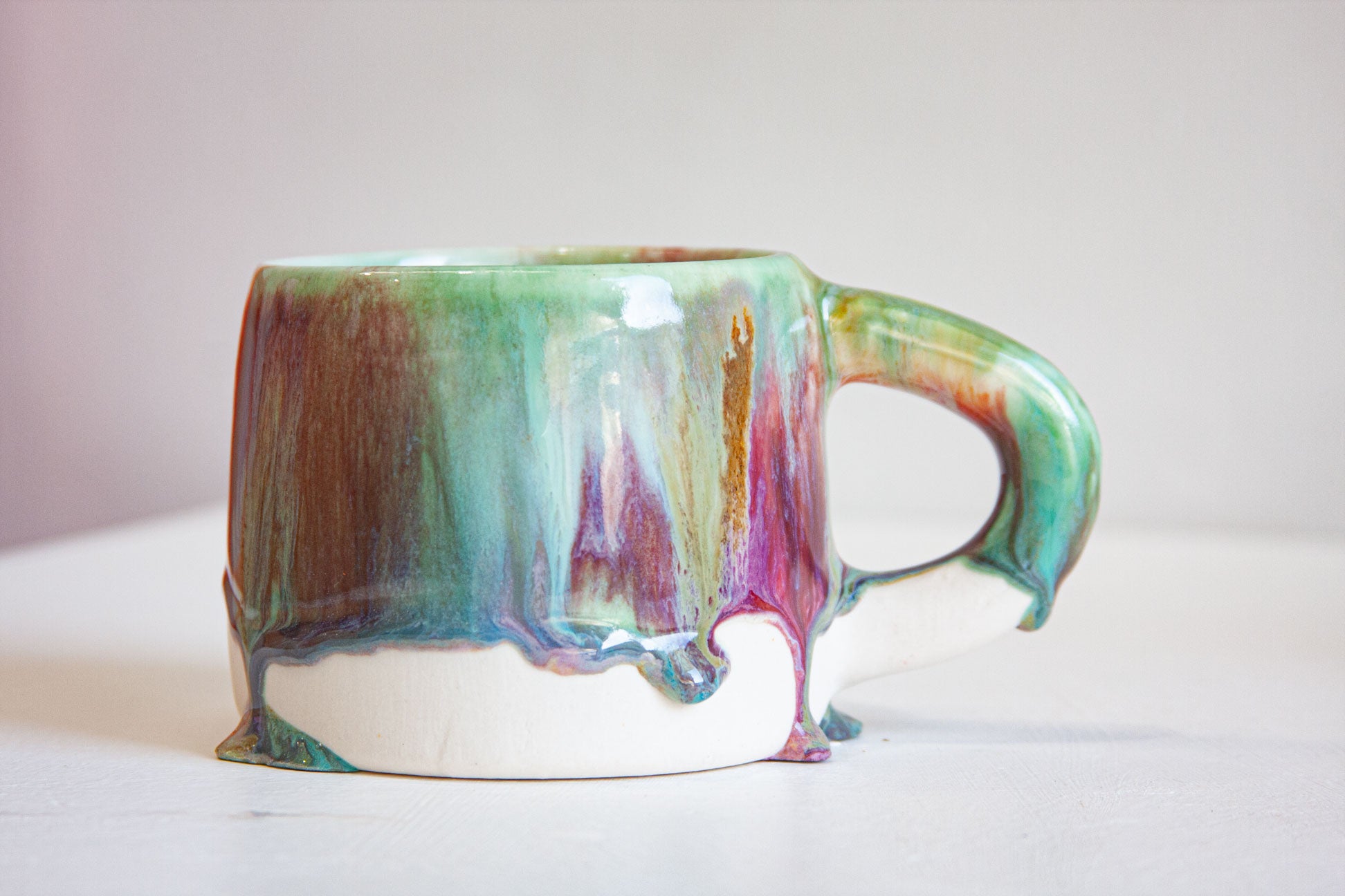 A ceramic mug with a dripping glaze effect, featuring a white base and a top surface decorated with a multicolor blend of green, teal, purple, and brown, set against a plain light background.