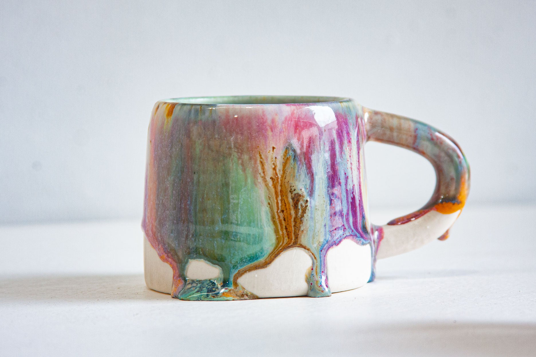 A single handmade ceramic mug seen side on, the green, pink and peach glaze streaming down the body and hanging in thick drips above the bare clay.