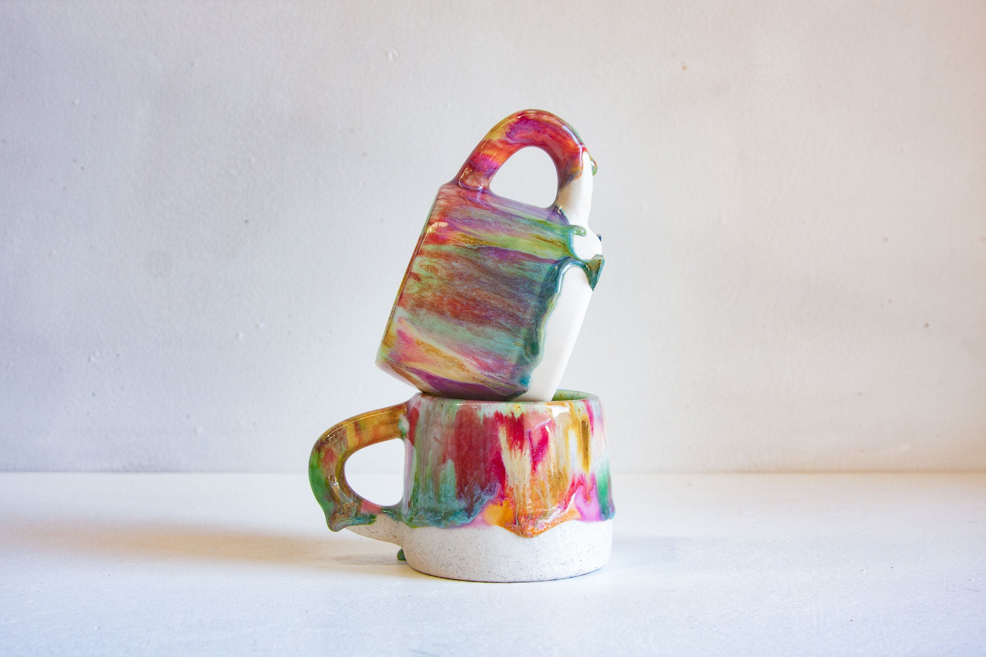 A stack of three small ceramic mugs with colorful marbled paint designs in pink, green, yellow, and blue on a white base, set against a plain light gray background.
