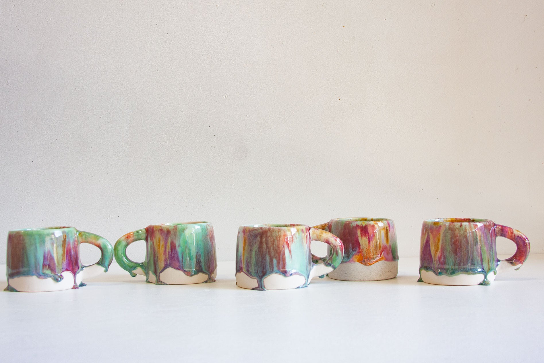 Five small ceramic mugs with colorful dripping glaze patterns in green, purple, orange, and pink, arranged in a row against a plain off-white background.