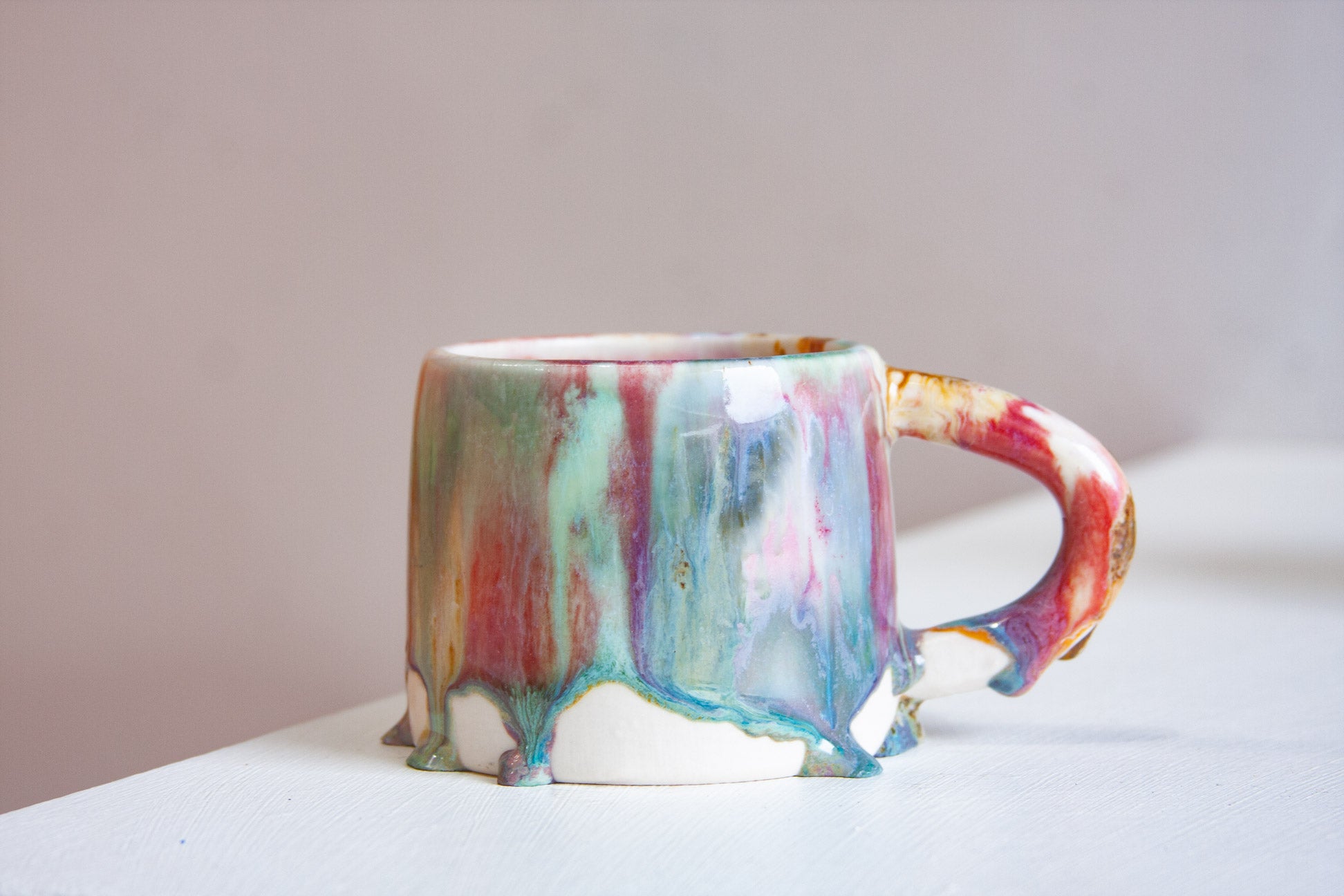 A handmade ceramic mug seen side on, the drips running down the body and over the handle in green, pink, peach and brown.