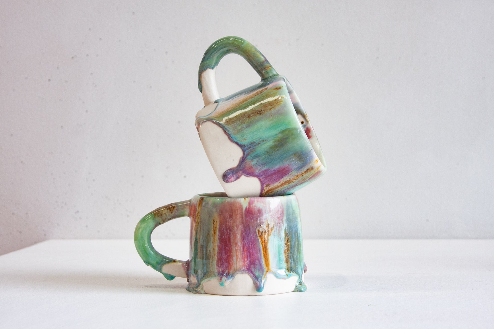Two handmade ceramic mugs stacked, one tilted on top of the other, showing the heavy drips running over the bare white clay bases.