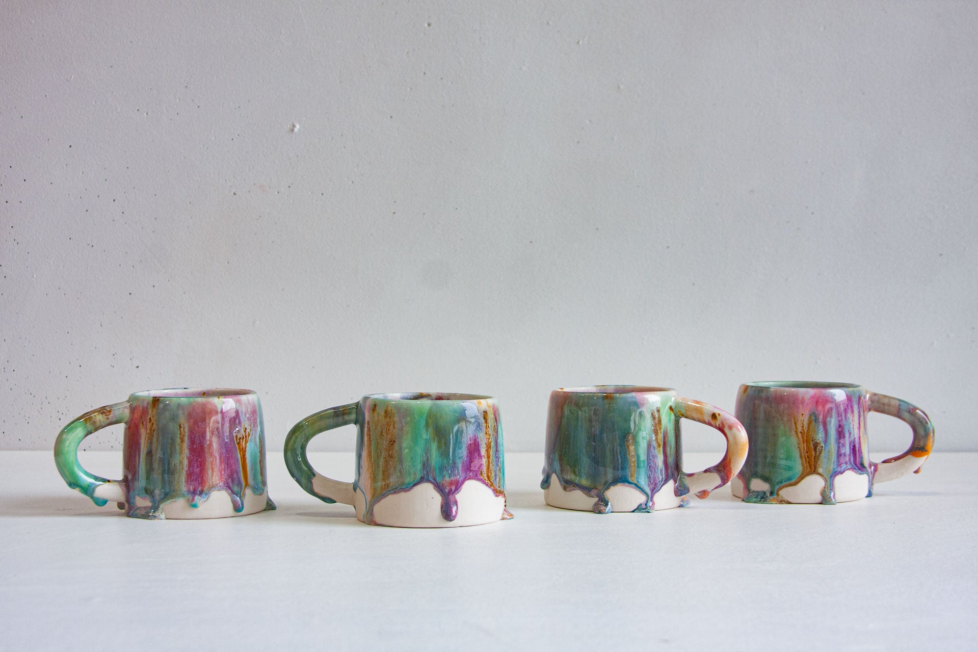 Four handmade ceramic mugs in a row, brush-glazed in green, pink, peach, brown and blue, the glaze running in heavy drips down to the bare white clay base.