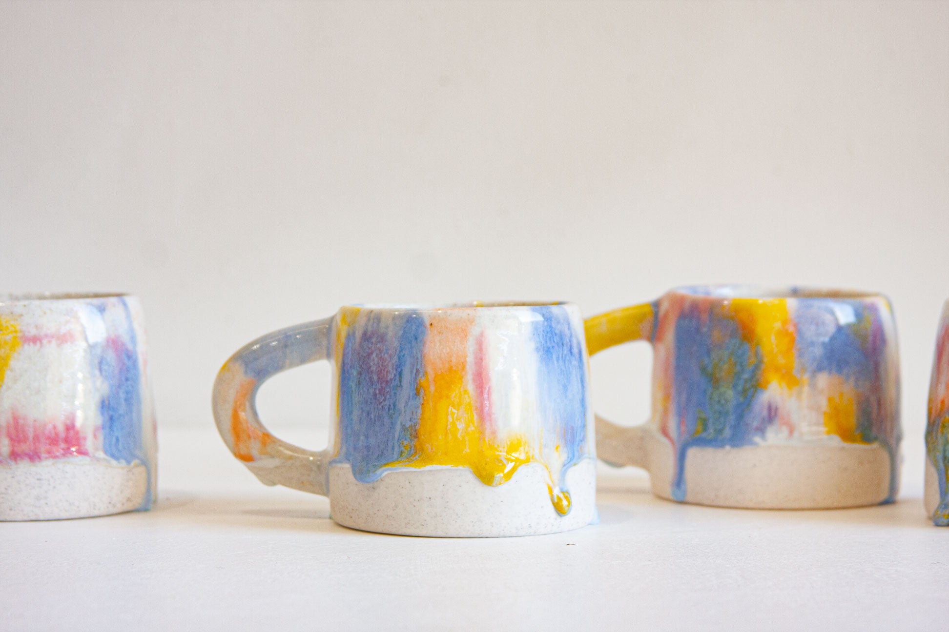 Three ceramic mugs with colorful dripping paint patterns in blue, yellow, pink, and orange on a white speckled base, arranged on a light surface against a plain off-white background.