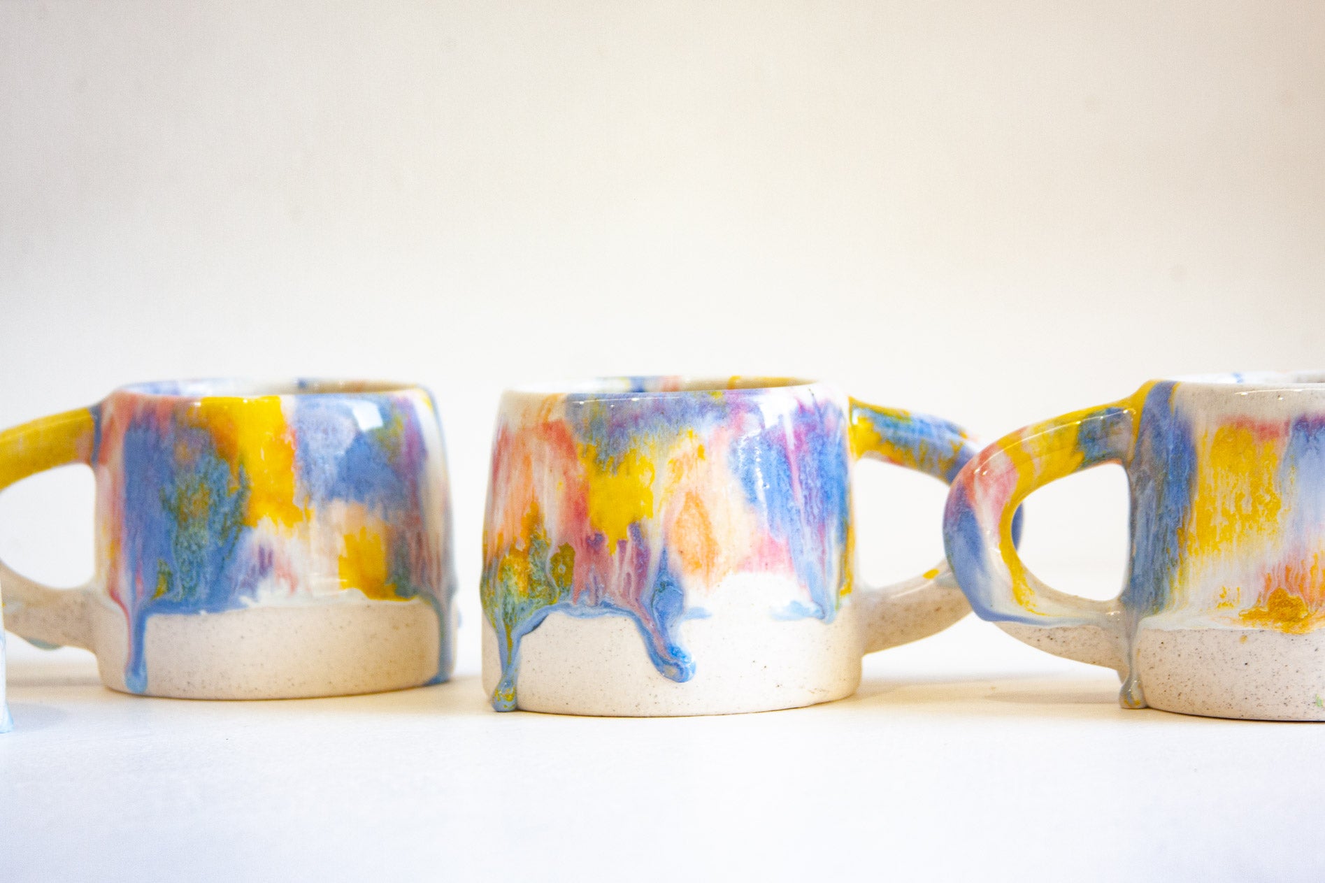 Three handmade ceramic mugs with colorful dripping glaze patterns in blue, yellow, pink, and purple on a white base, set against a plain off-white background.