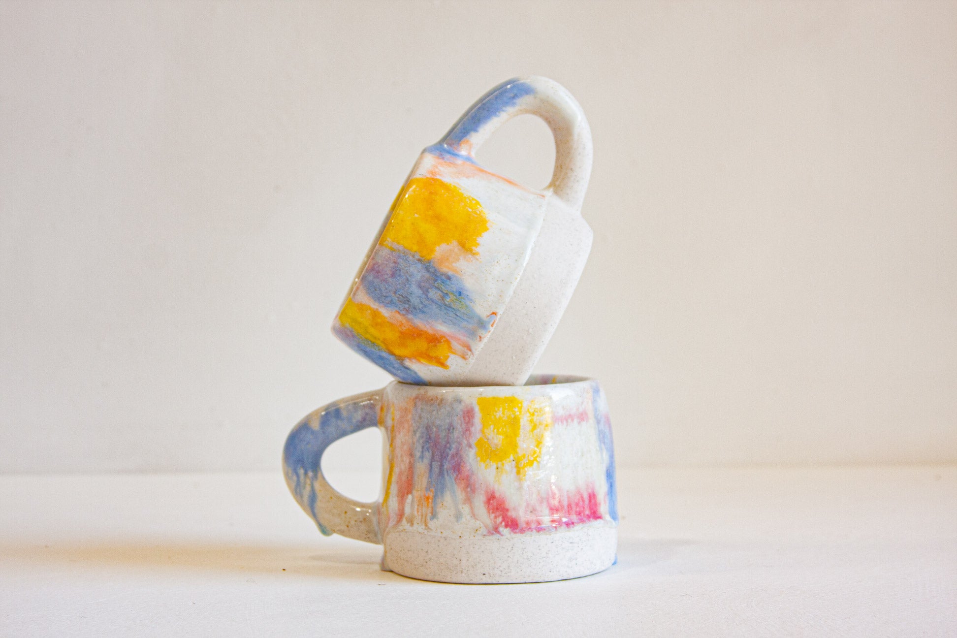Two small ceramic mugs with abstract painted patterns in yellow, blue, pink, and white, stacked with one tilted on top of the other against a plain light background.