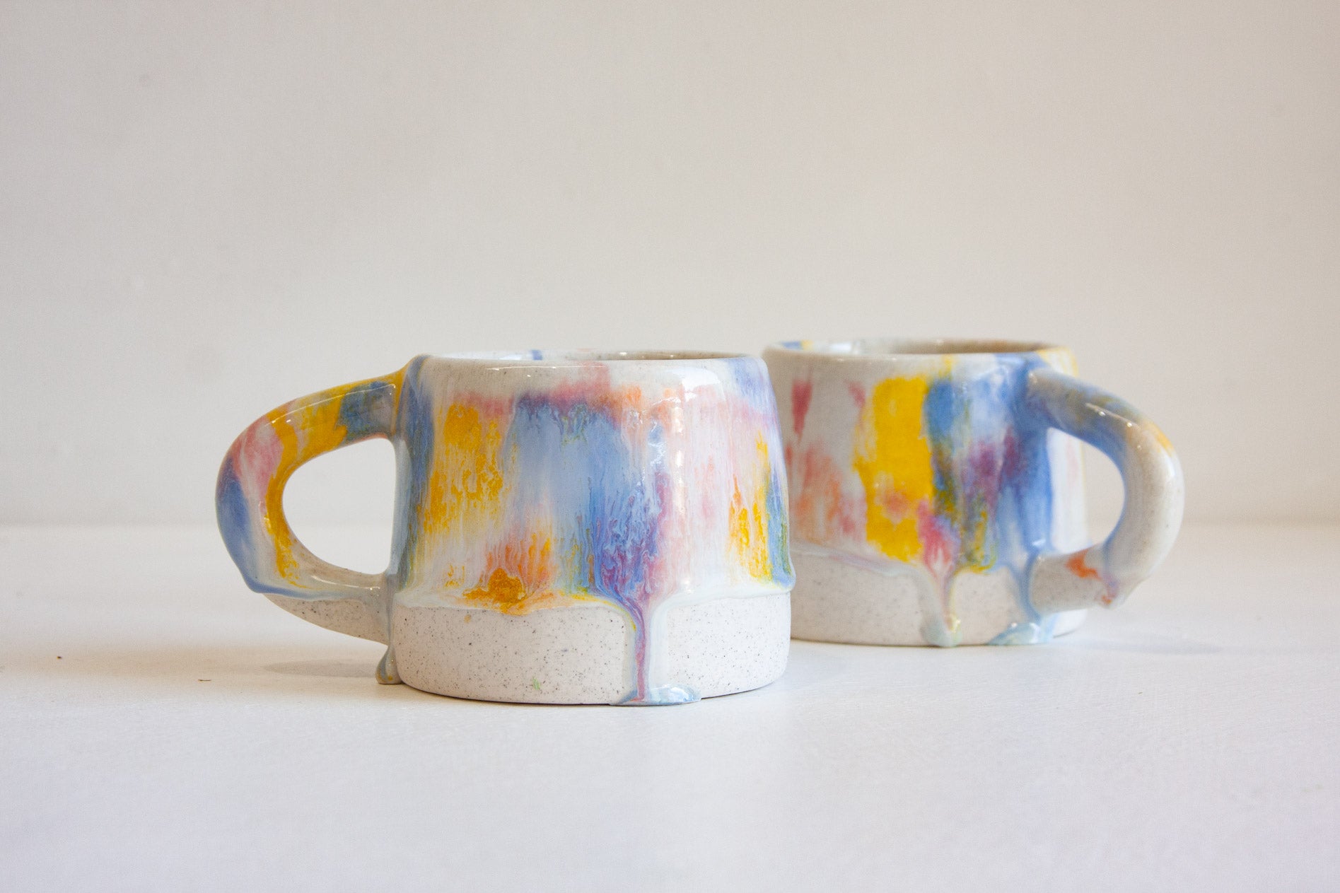 Two ceramic mugs with dripping multicolor glaze, featuring blue, yellow, pink, and orange paint-like drips on a white base, set against a plain white background.