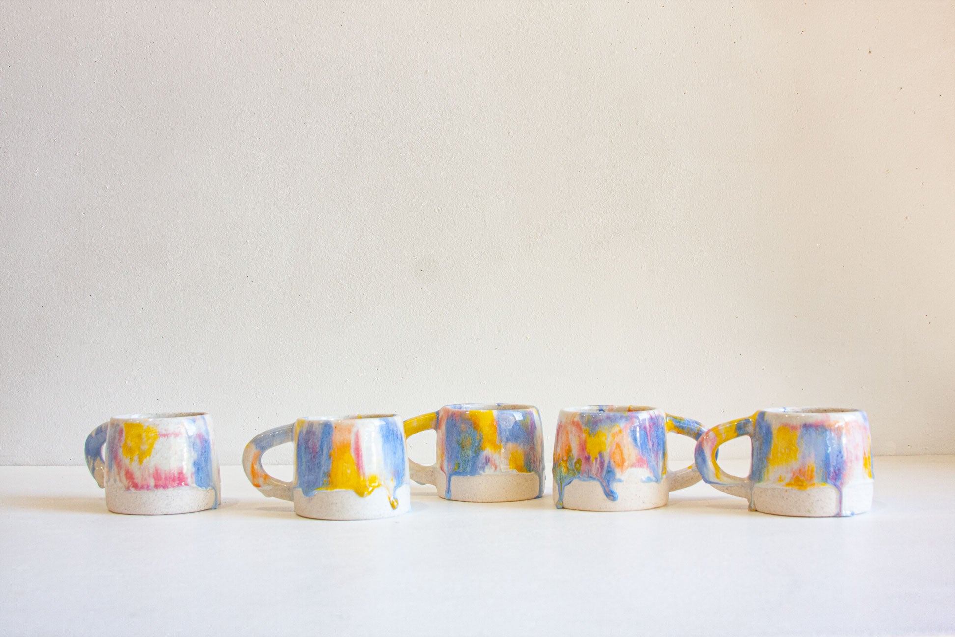 Five small ceramic mugs with colorful abstract paint splatter patterns in blue, yellow, pink, and white, lined up on a white surface against a plain beige wall.