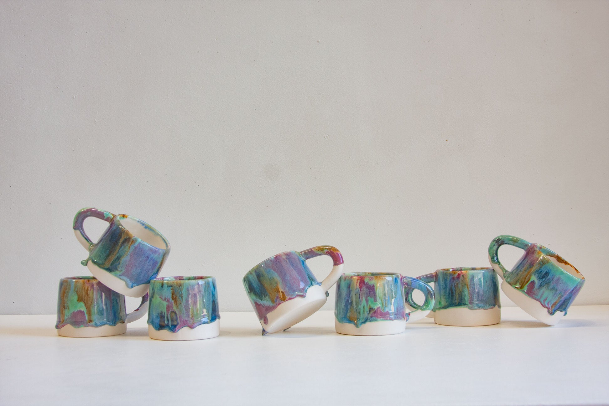 Seven ceramic coffee mugs with colorful dripping glazes in shades of blue, green, purple, and gold, arranged on a white surface against a plain beige background.