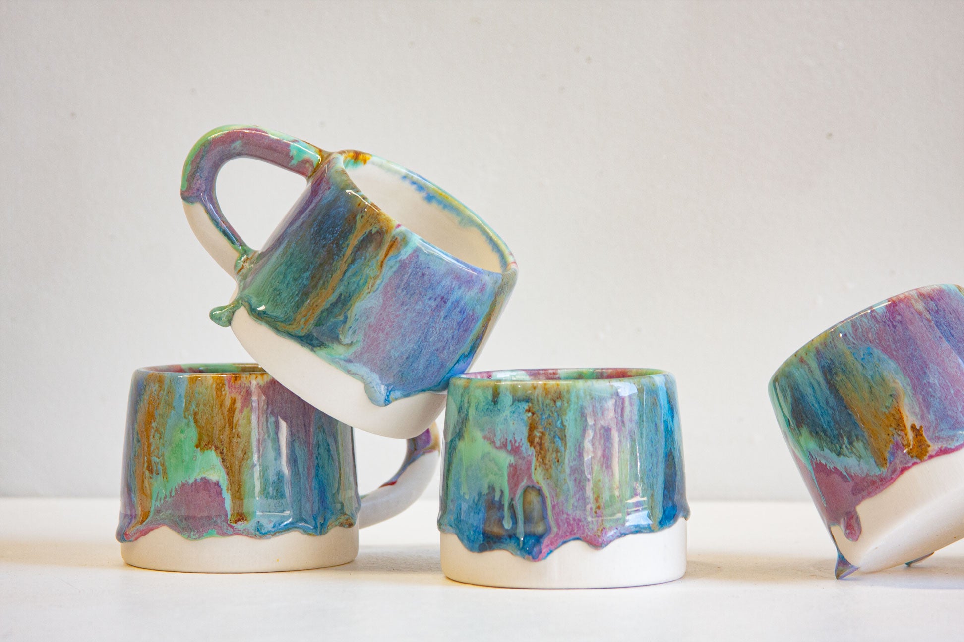 Three handmade ceramic mugs with dripping multicolor glaze, displayed on a white surface against a light beige background.