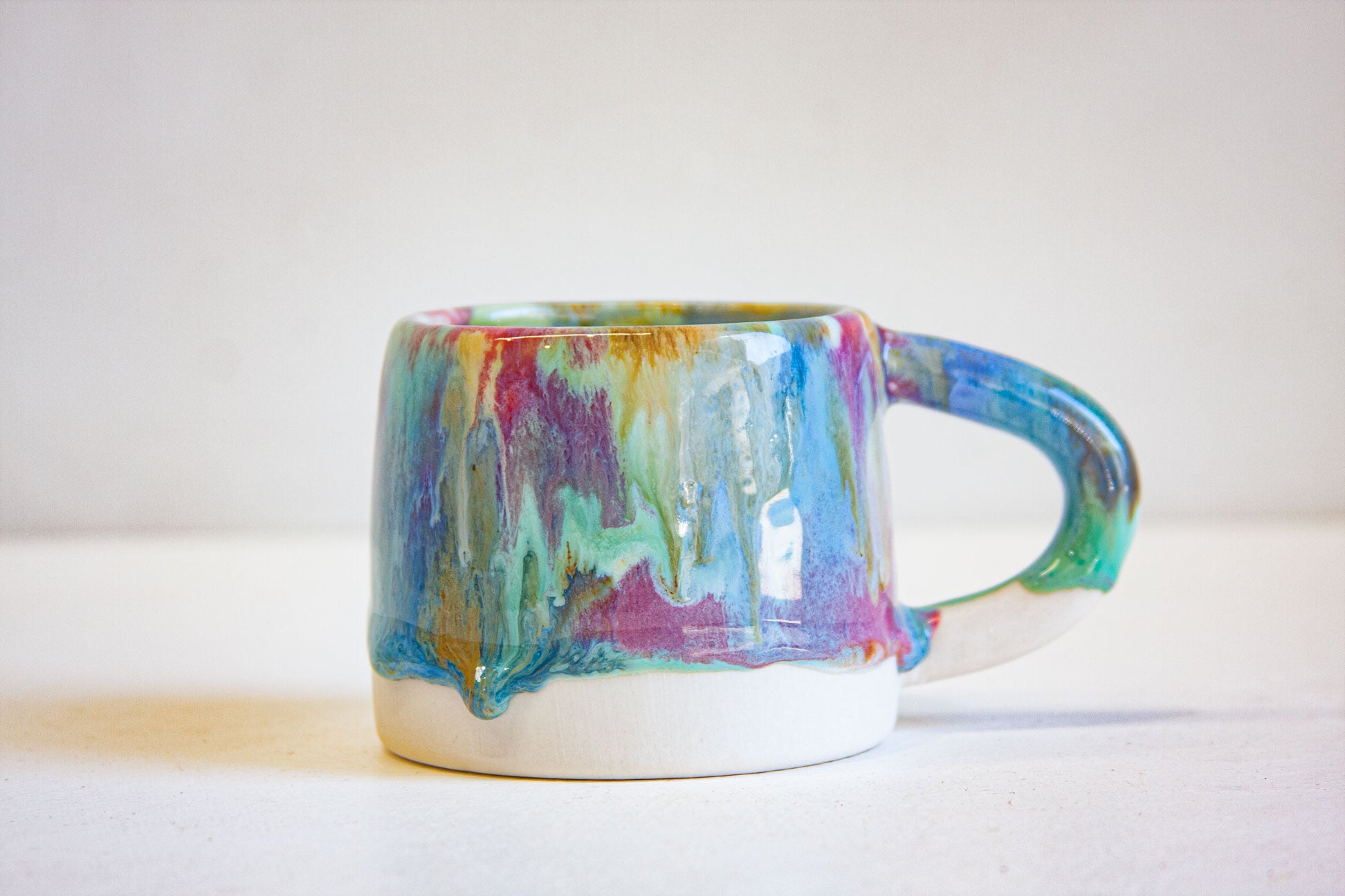 A handmade ceramic mug with a dripping glaze effect, featuring vibrant colors of blue, green, purple, and gold on the upper portion, with a solid white base and a matching curved handle.