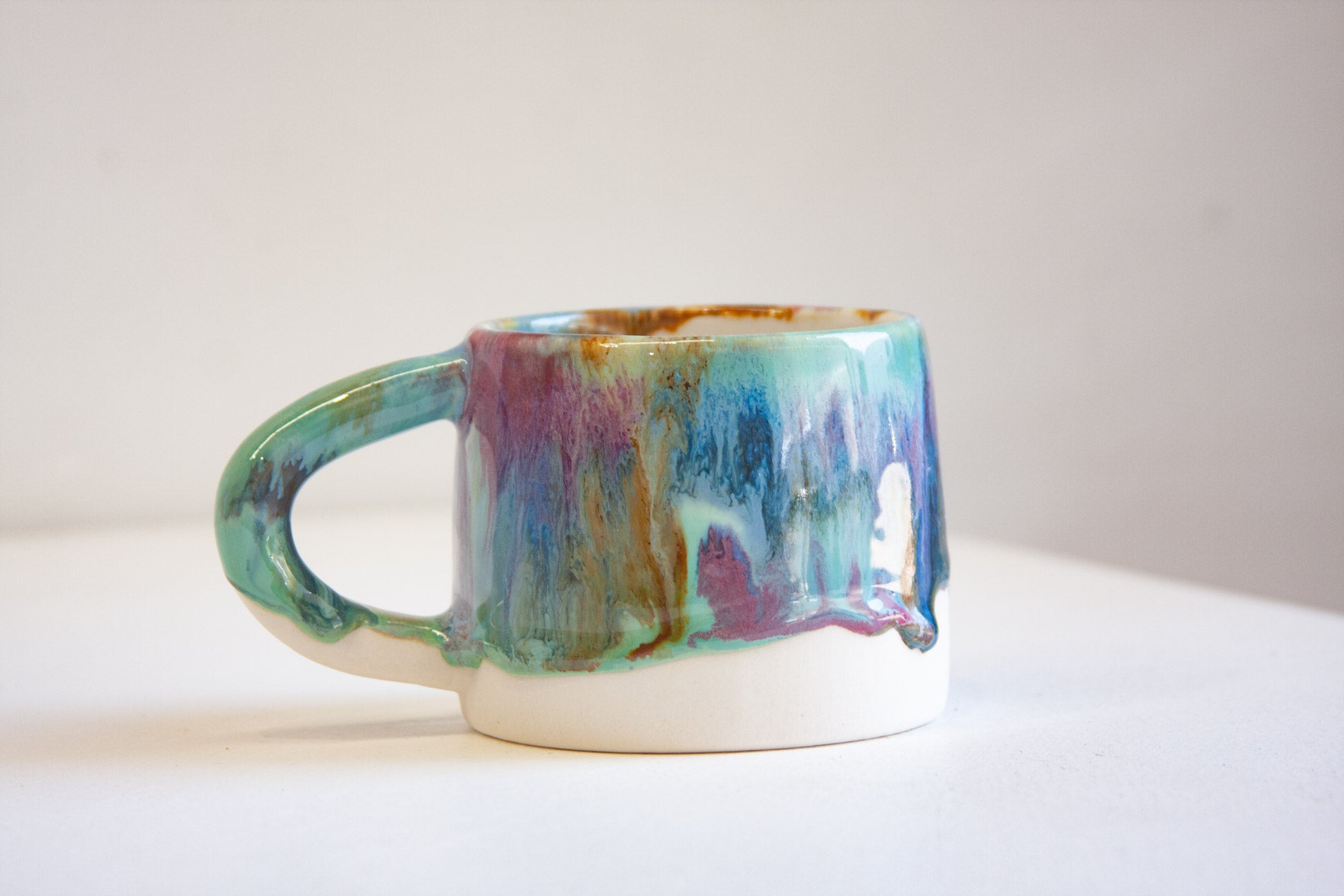 A ceramic mug with a dripping glaze design featuring teal, purple, blue, and brown on a white base, set against a plain light background.
