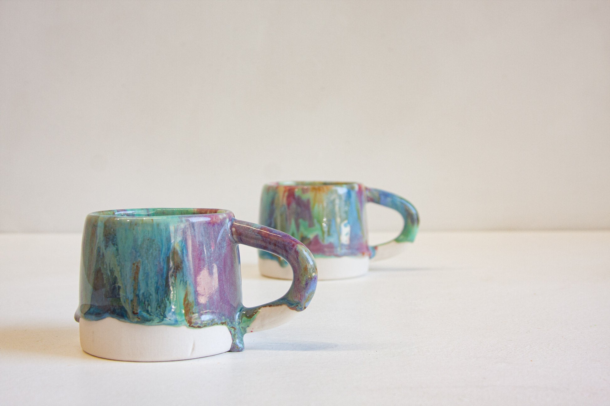 Two small ceramic mugs with a colorful, marbled glaze in shades of teal, green, purple, and blue, sitting on a white surface against a light beige background.