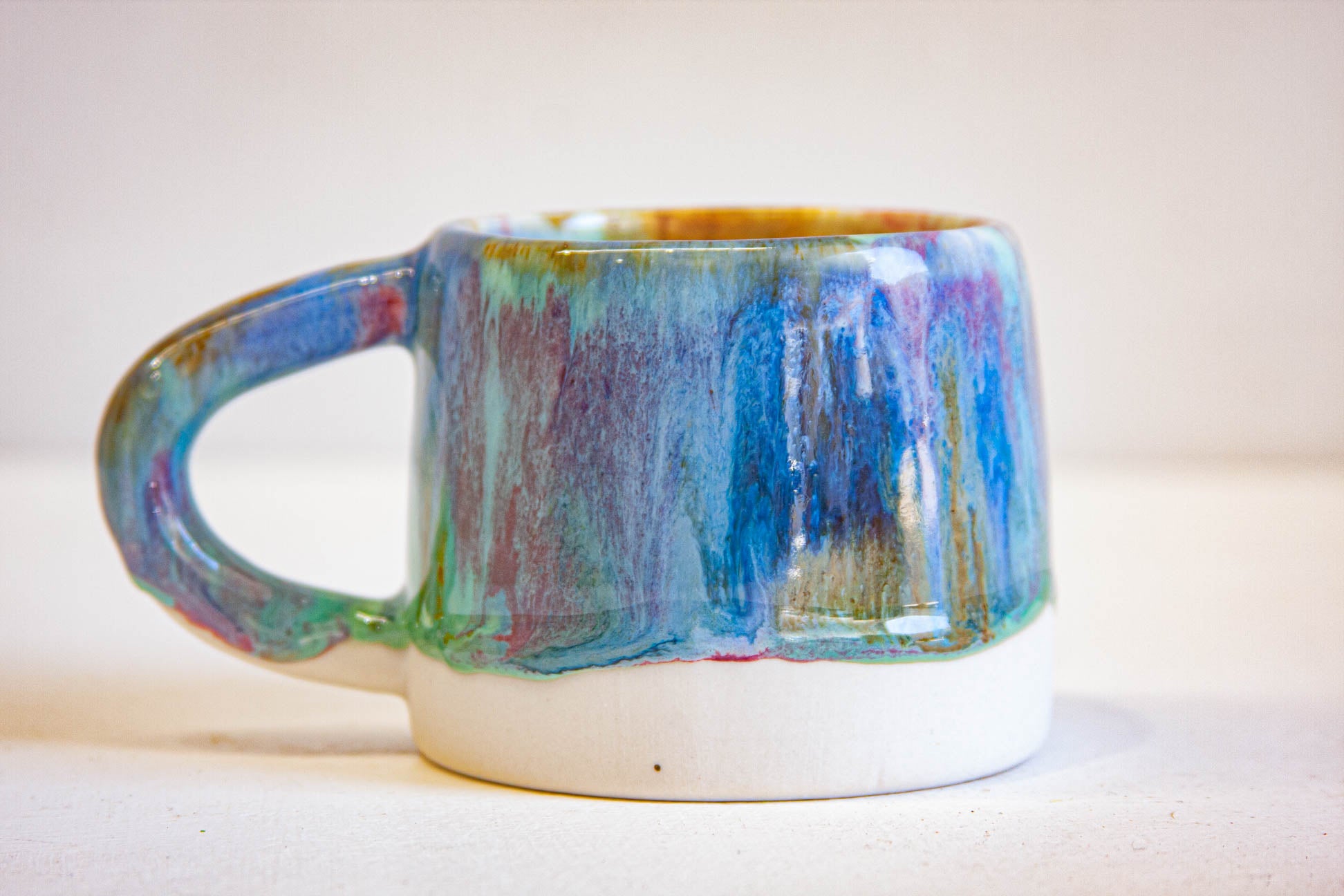 A small ceramic mug with a glossy, multicolored dripping glaze in shades of blue, green, purple, and gold, featuring a solid white base.