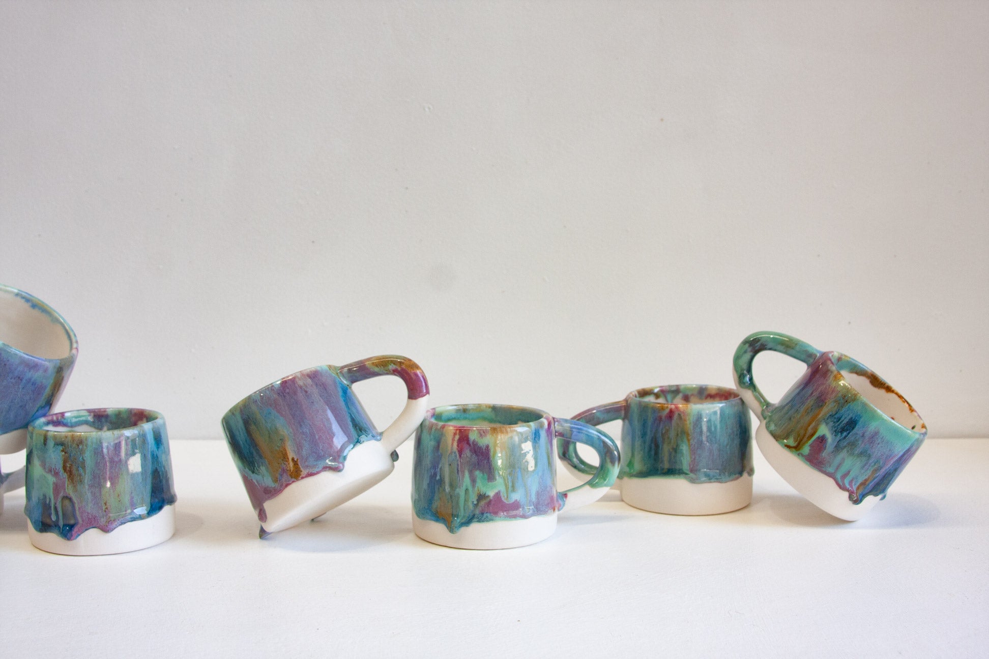 A set of handmade ceramic mugs with colorful, dripping glaze in shades of teal, blue, purple, and green, displayed on a white surface against a light gray background.