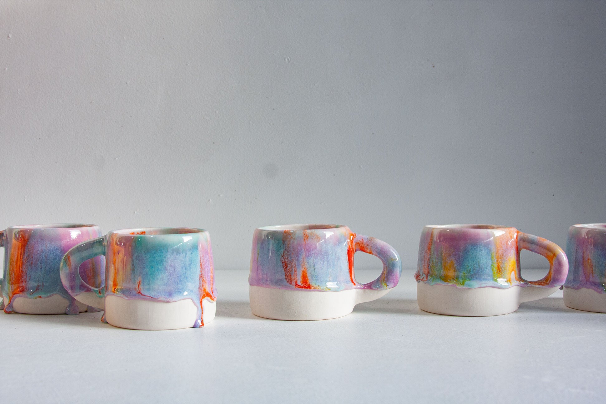 Five handmade ceramic mugs arranged in a row, glazed in teal, lilac and orange drips over a white lower half, against a plain pale background.