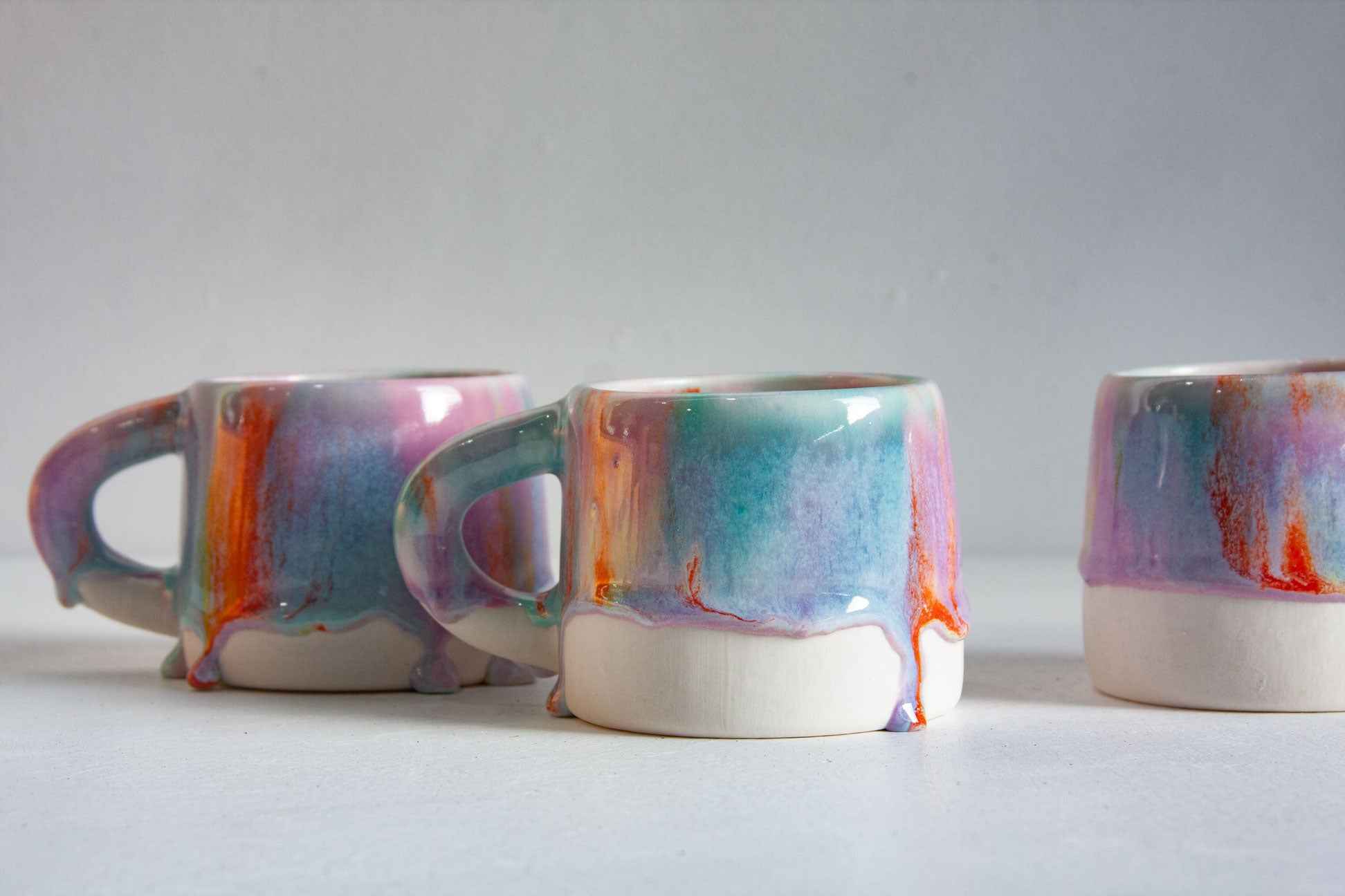 Three handmade ceramic mugs seen close up, glazed in teal, blue and lilac with bright orange drips running over the white lower half.