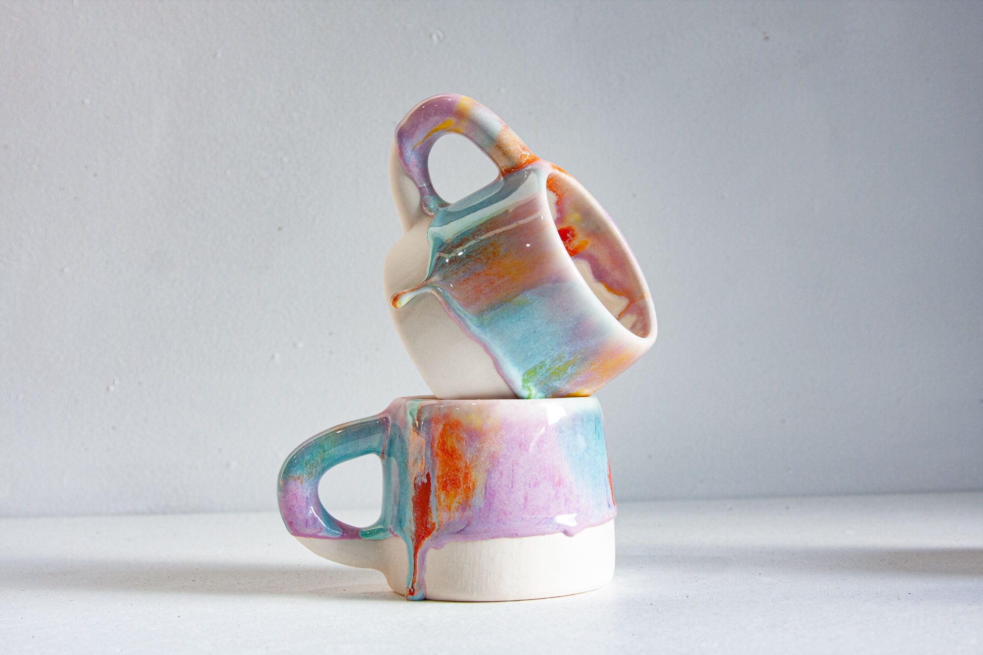 Two handmade ceramic mugs stacked one on the other, glazed in lilac, teal, orange and yellow drips running over a white lower half, against a pale grey wall.