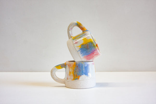 Two handmade ceramic mugs stacked at an angle, multicolour blue, yellow and pink drip glaze over unglazed speckled white stoneware base, curved handles