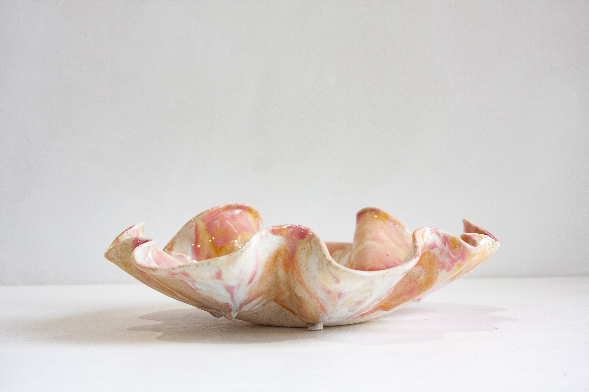 A handmade ceramic oyster platter seen from the side, the folds of the rim curving up and outward, glazed in pink, peach and cream.