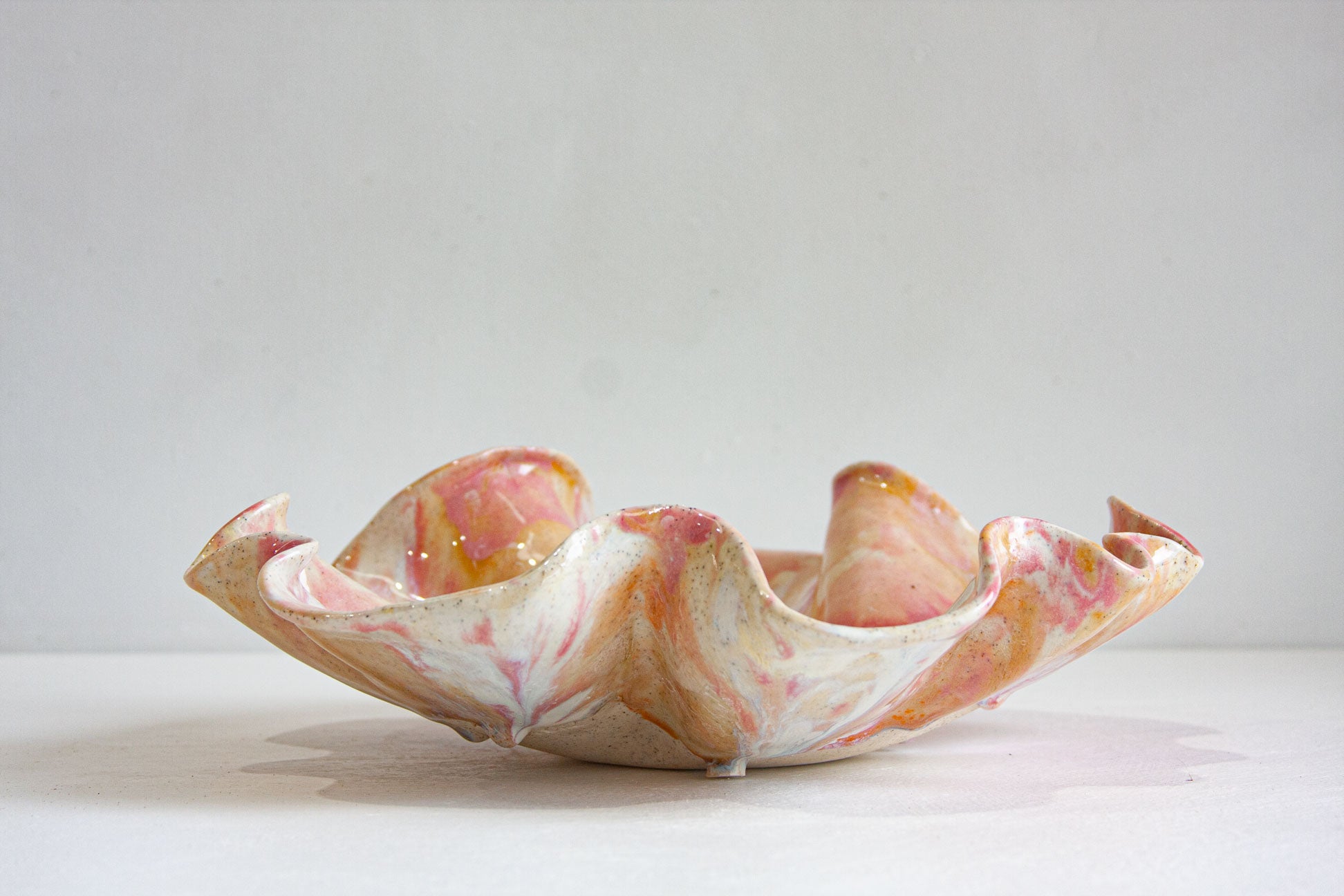 A handmade ceramic oyster platter with a sculptural wavy rim, glazed in soft pink, peach and cream over speckled clay, on a white surface against a pale wall.