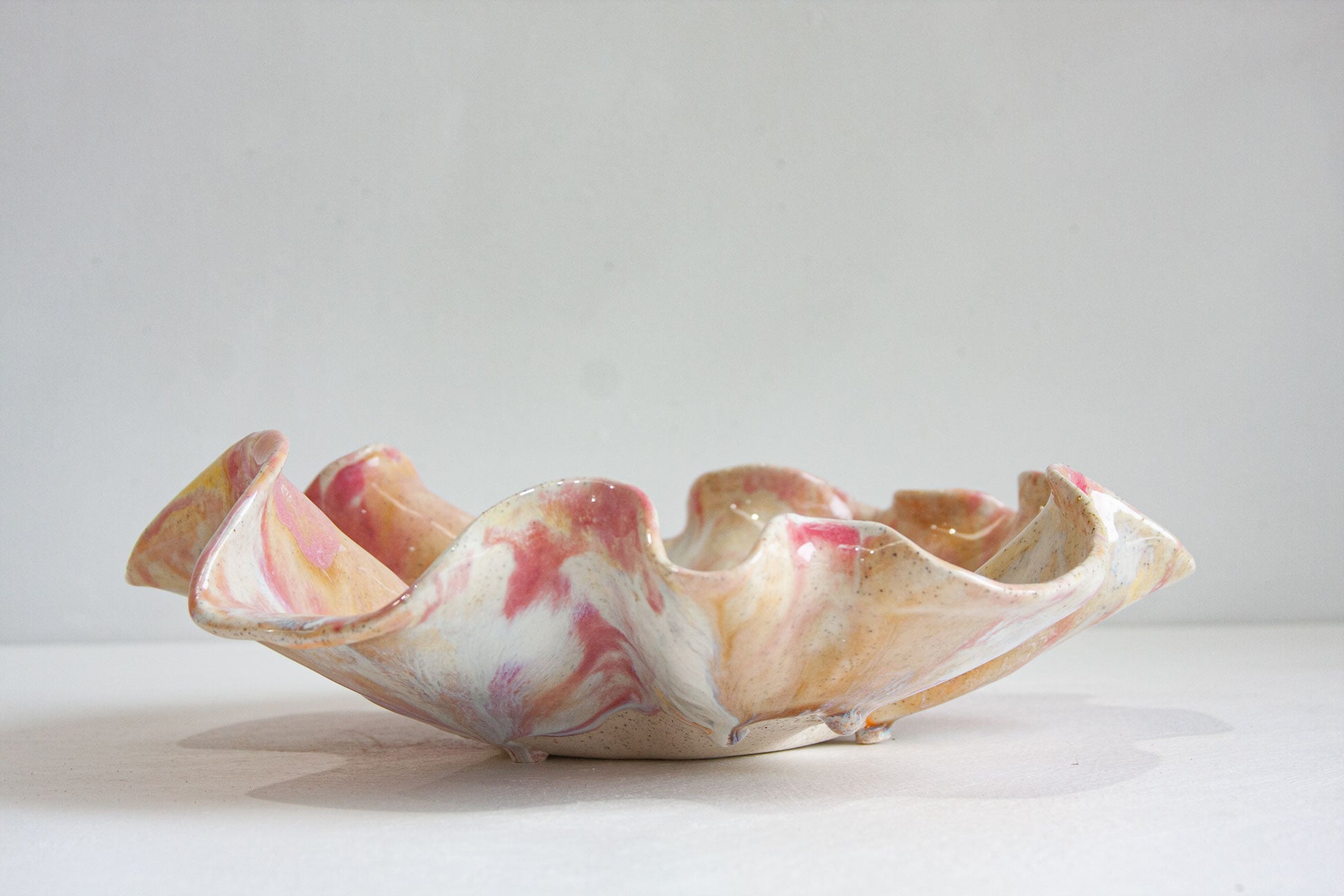 A handmade ceramic oyster platter seen from the side, its folded rim rising into soft peaks, glazed in pink, peach and cream over speckled clay.