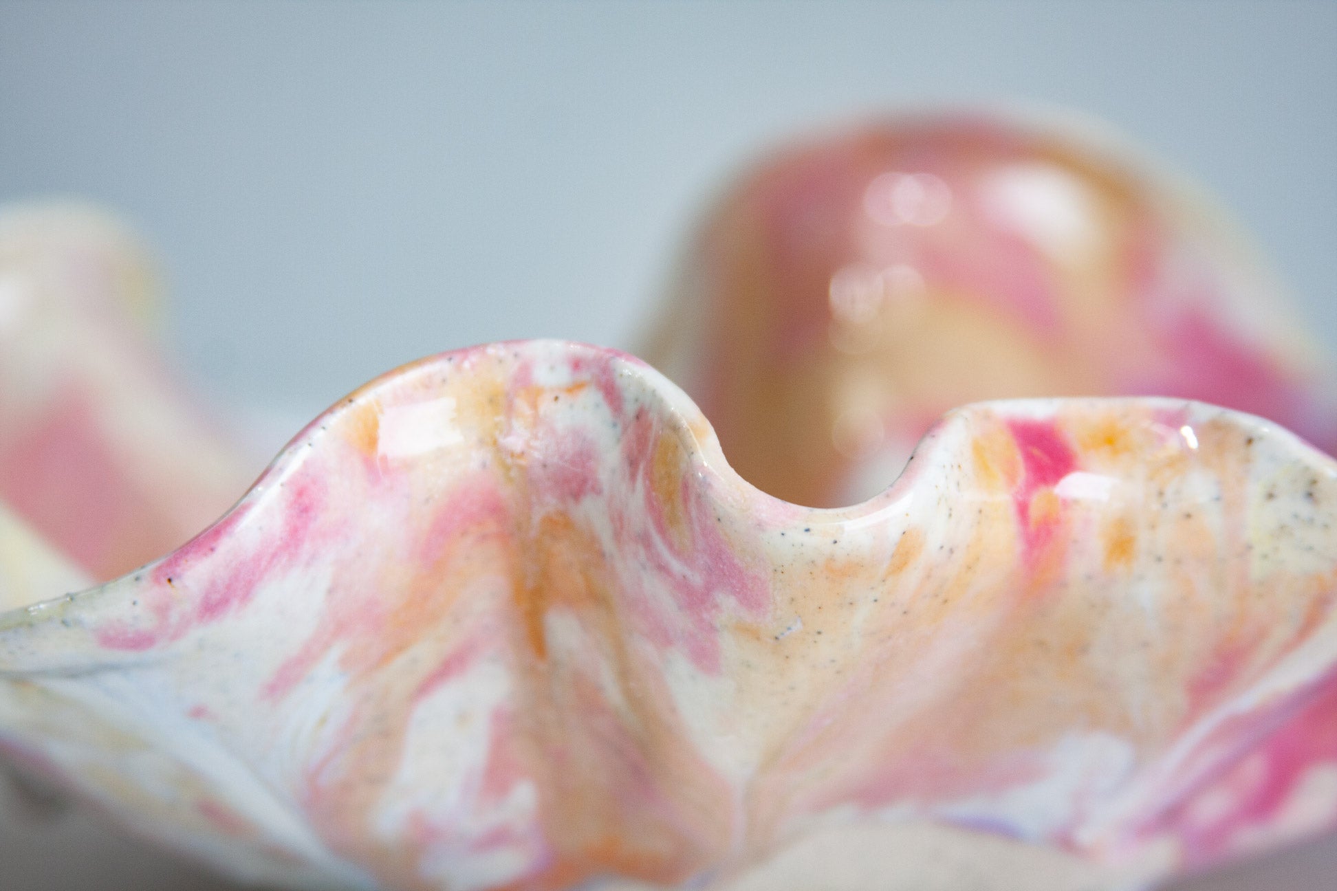 Close-up of the rim of a handmade ceramic oyster platter, pink and orange running through cream over speckled clay, with the folds curving out of focus behind.