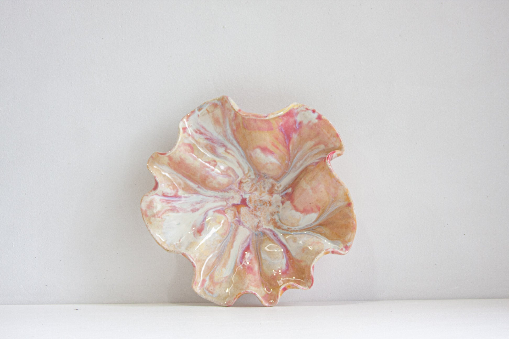 A handmade ceramic oyster platter stood upright to show its face, glazed in pink, peach and cream over speckled clay, against a pale wall.