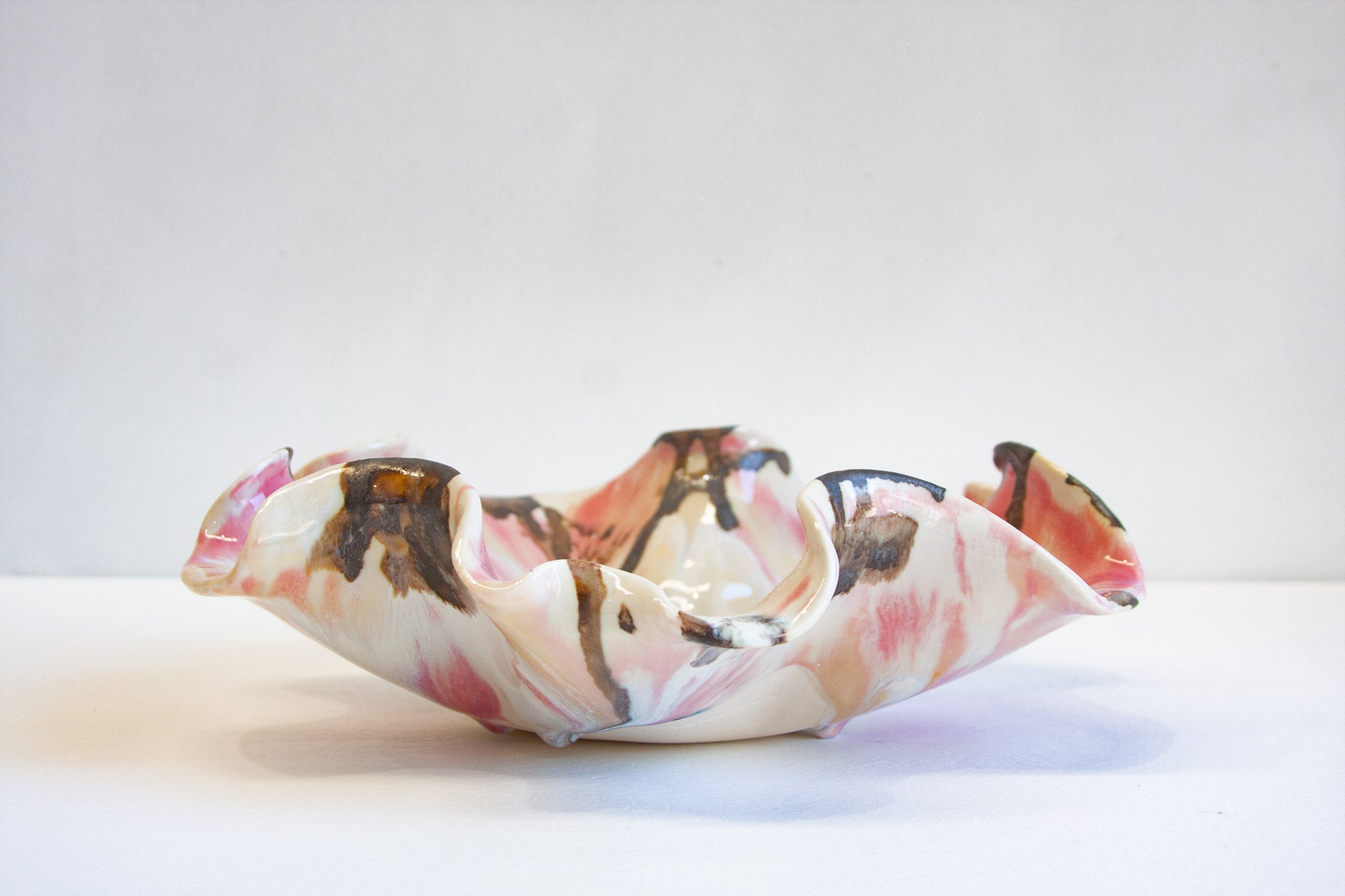 A handmade ceramic bowl with a ruffled, organic edge, decorated with a marbled pattern of pink, brown, and cream on a white base, photographed on a plain white surface.