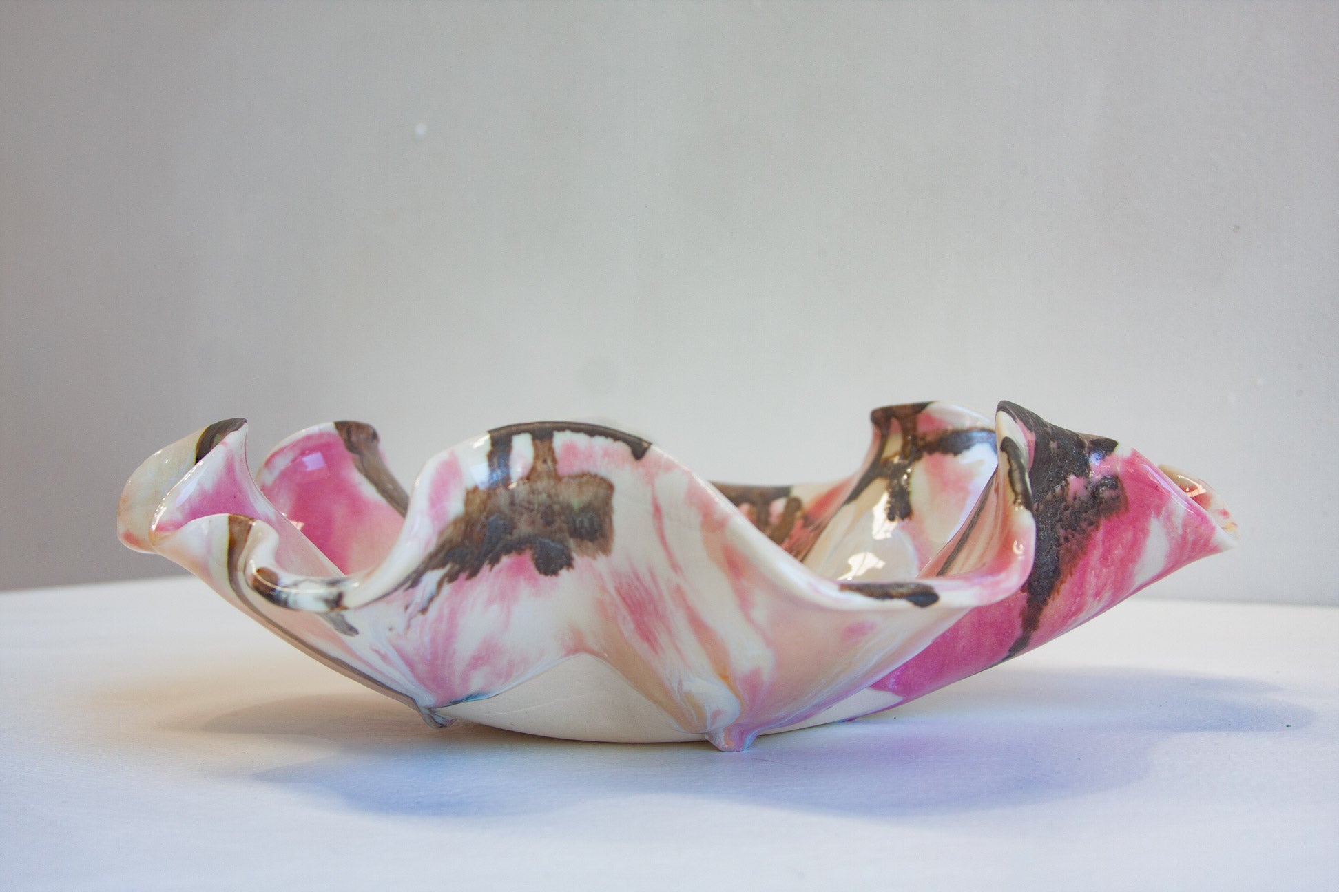 A handmade ceramic bowl with a ruffled, scalloped edge, featuring a marbled pattern of pink, white, and dark brown, displayed on a white surface against a neutral gray background.