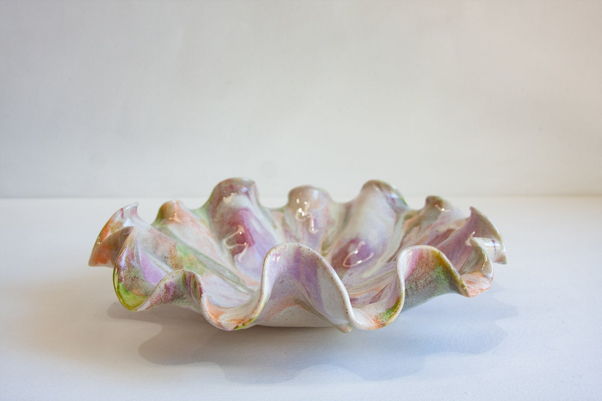 A decorative ceramic bowl with a ruffled, scalloped edge, featuring a marbled glaze of white, lavender purple, pink, and green, set against a plain light background.