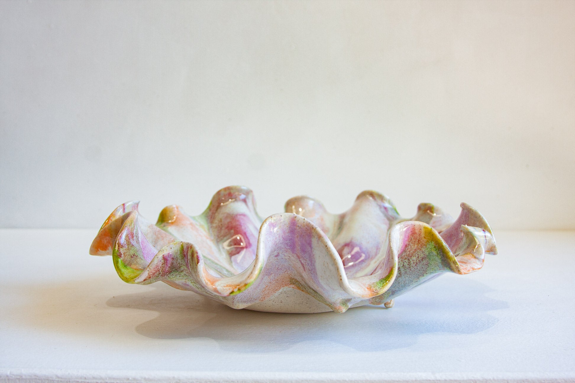 A decorative ceramic bowl with a ruffled, scalloped edge, featuring a glossy glaze in pastel pink, lavender, green, and cream tones, displayed on a white surface against a neutral light gray background.