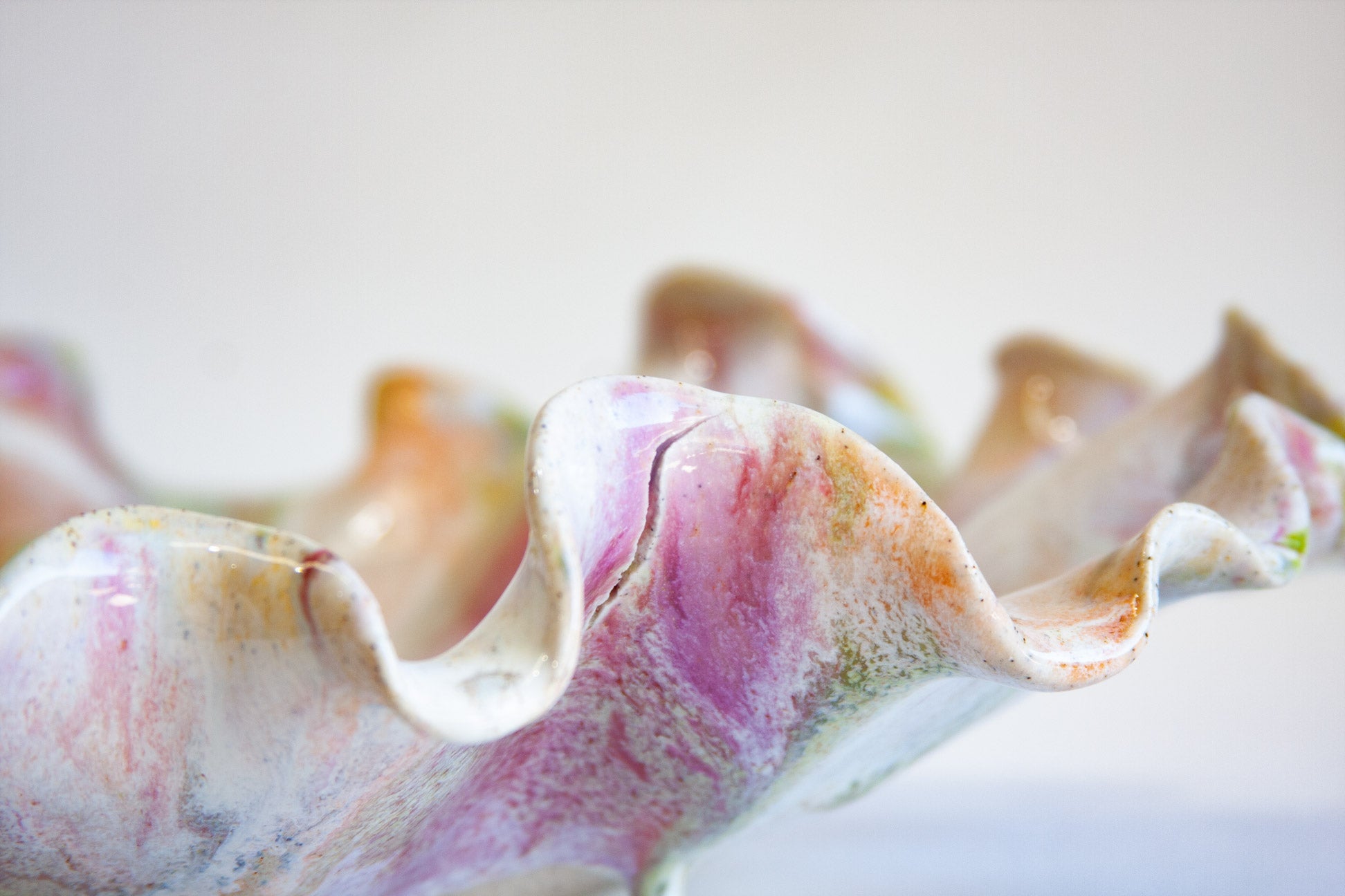 A close-up of a decorative ceramic shell-shaped vessel with wavy ruffled edges, featuring a marbled glaze in soft pink, purple, beige, and white tones.