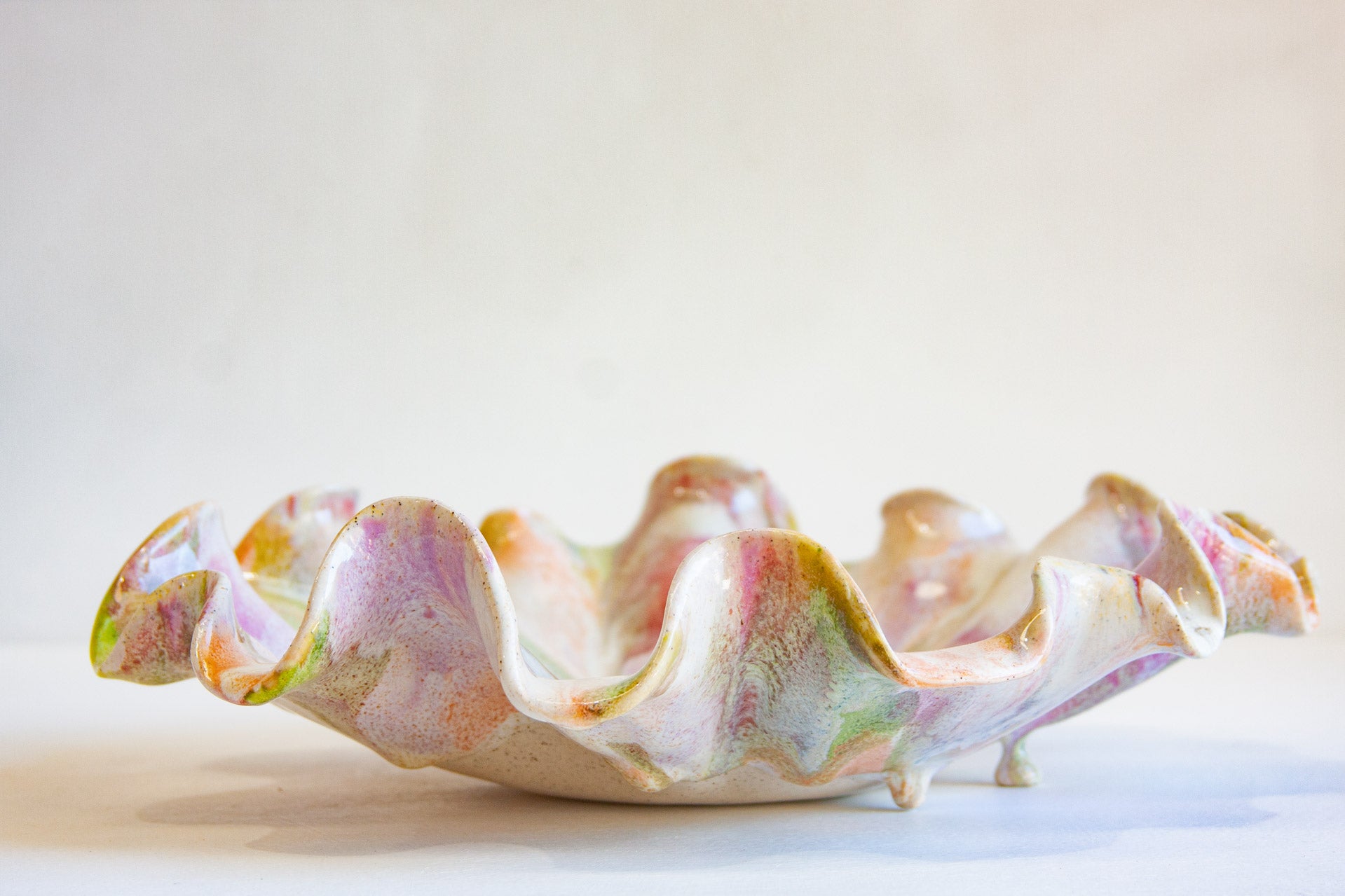 A handmade ceramic bowl with a wavy, ruffled edge and small feet, decorated with a marbled glaze of pink, green, orange, and cream, set against a plain light background.