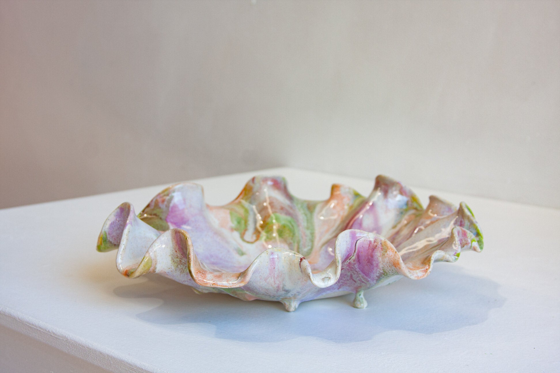 A ruffled ceramic bowl with a glossy finish, decorated with pastel pink, purple, green, and orange marbled patterns on an off-white base, displayed on a white surface against a neutral beige background.