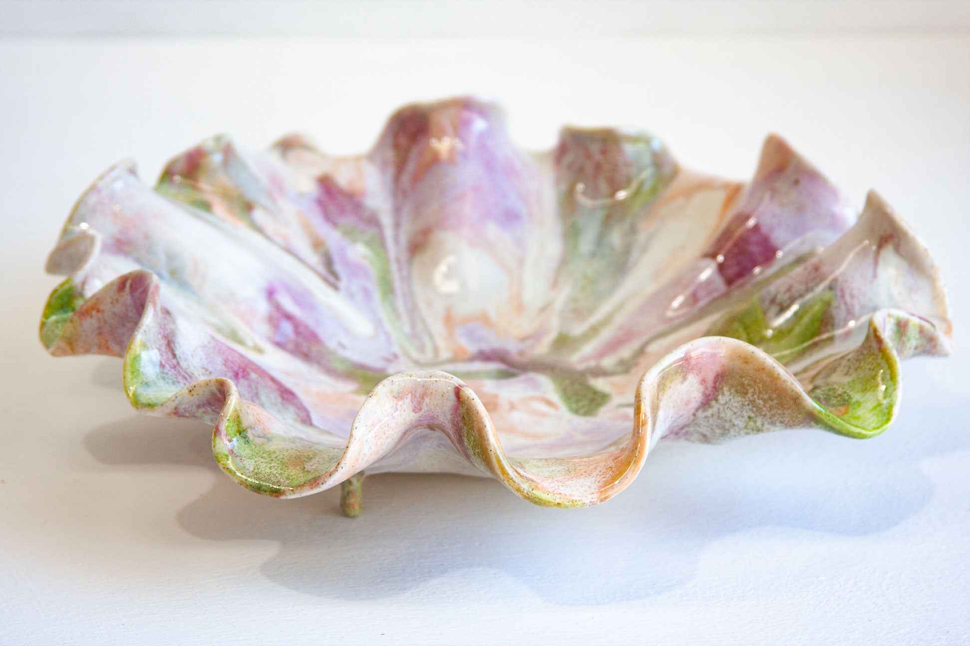 A handmade ceramic dish with a ruffled, petal-like edge, decorated with a marbled glaze of pink, purple, green, and cream on a white background.