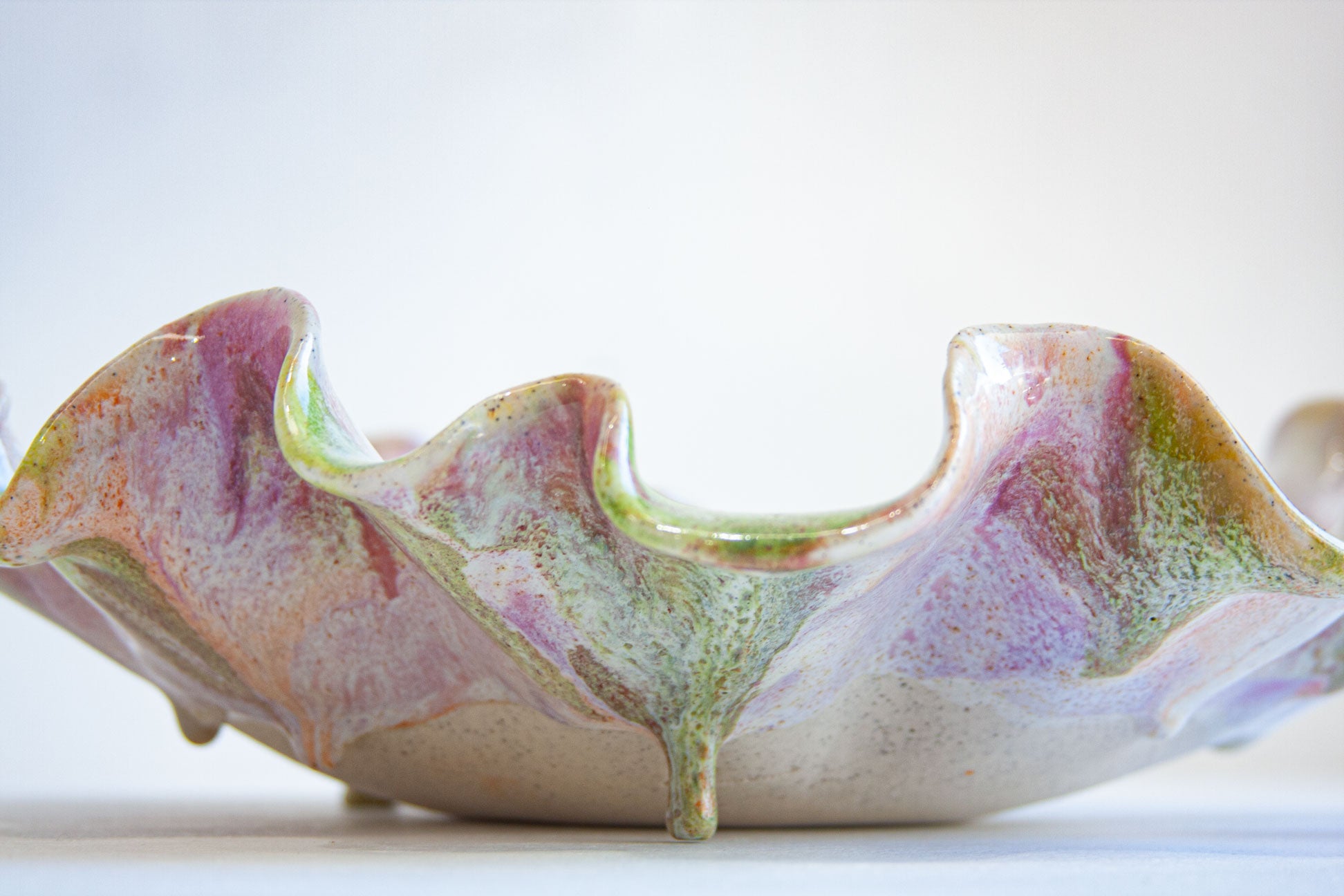 A close-up of a sculptural ceramic bowl with a wavy, organic rim, featuring a marbled glaze of pink, purple, green, and beige on a white background.
