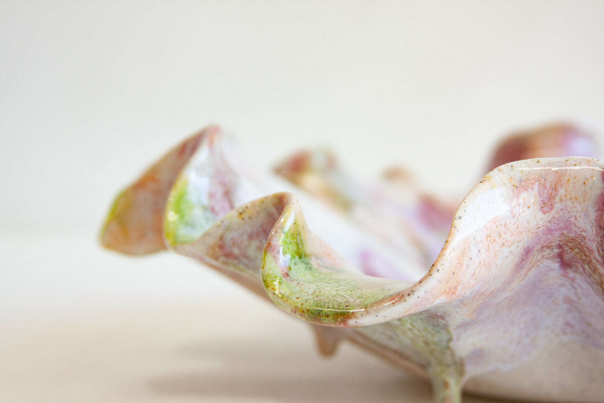 A close-up of a sculptural ceramic dish with a seashell-inspired wavy rim, featuring a marbled glaze of soft pink, lavender, green, and cream on a warm off-white background.