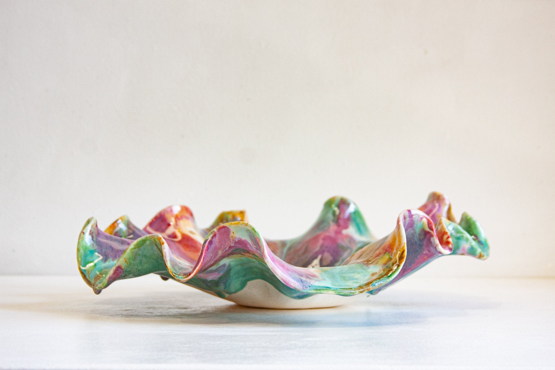 A handmade ceramic bowl with a wavy, ruffled rim, decorated in a marbled pattern of pink, green, blue, and purple glazes with gold accents, set against a plain off-white background.