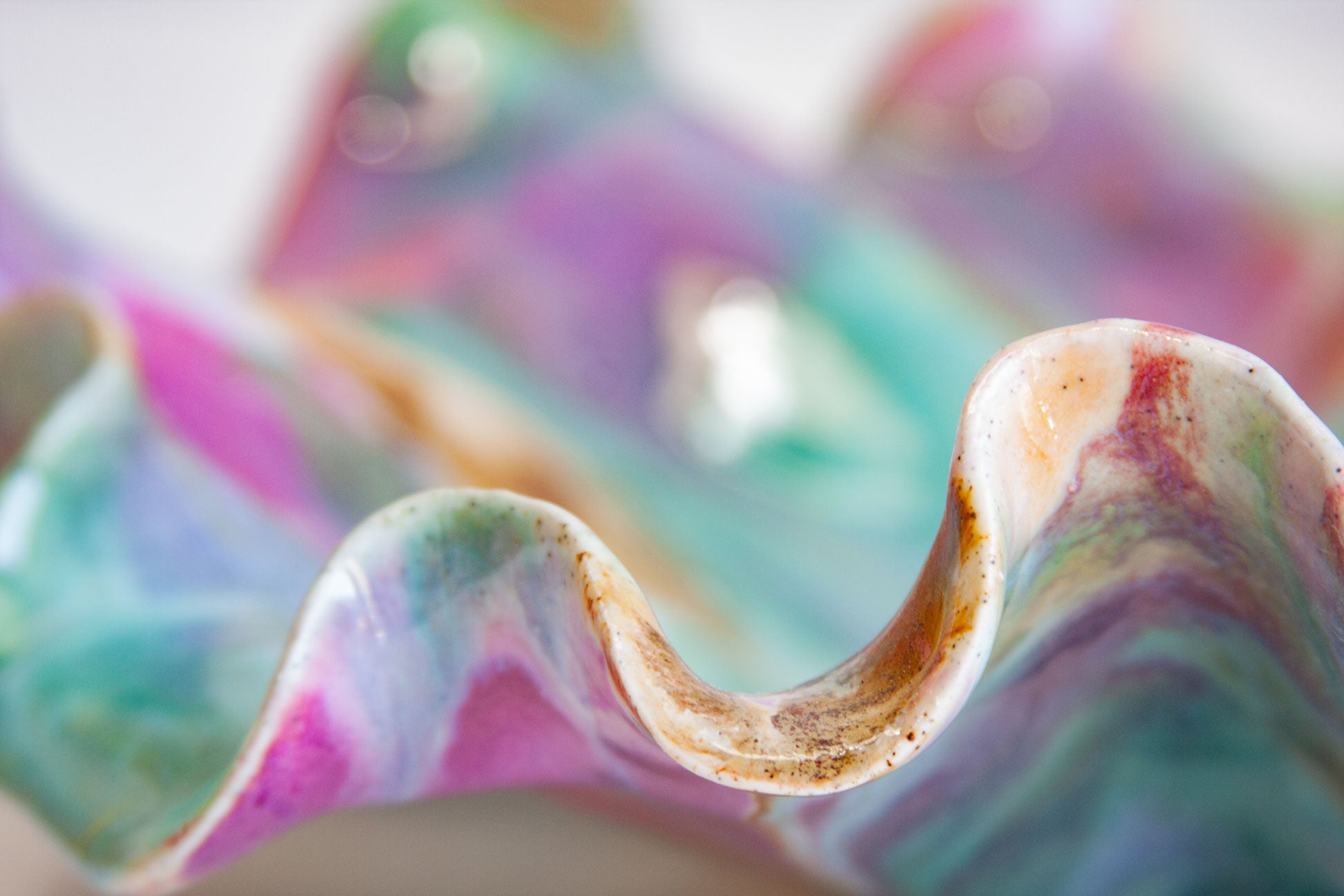 A close-up of a wavy, shell-like ceramic vessel with a glossy, marbled finish in pastel pink, purple, teal, and blue, with a small metallic silver accent visible inside.