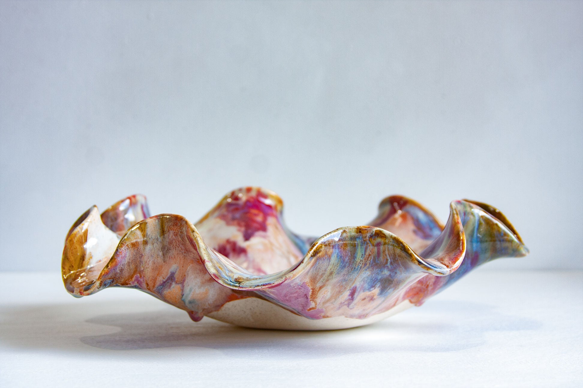 A handmade ceramic bowl with a wavy, ruffled edge, decorated with a marbled glaze of pink, purple, blue, and gold, set against a neutral light gray background.