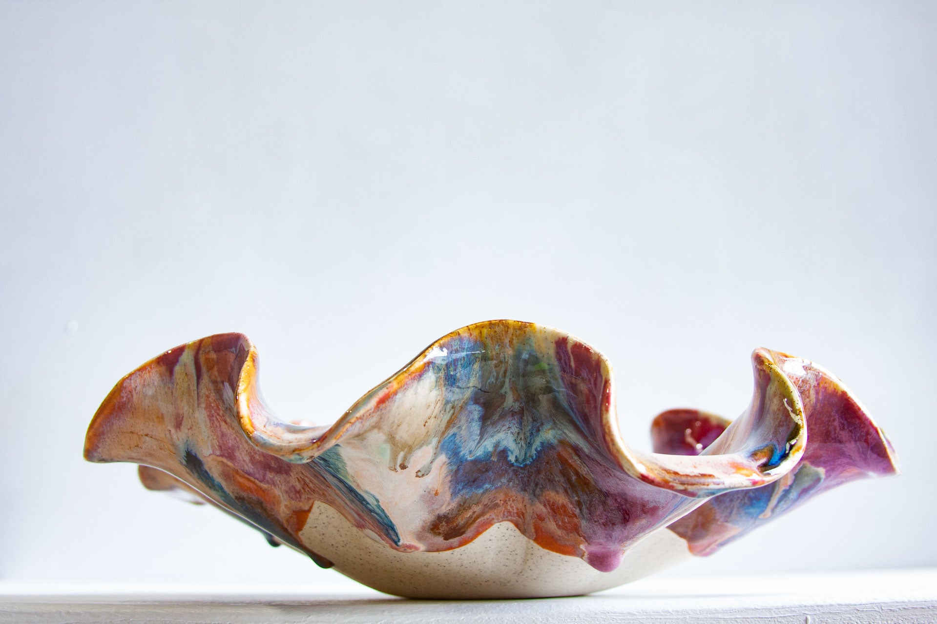 A handmade ceramic bowl with a wavy, flared rim, decorated with marbled, fluid glazes in shades of purple, blue, orange, and brown over a cream base, set against a plain light gray background.
